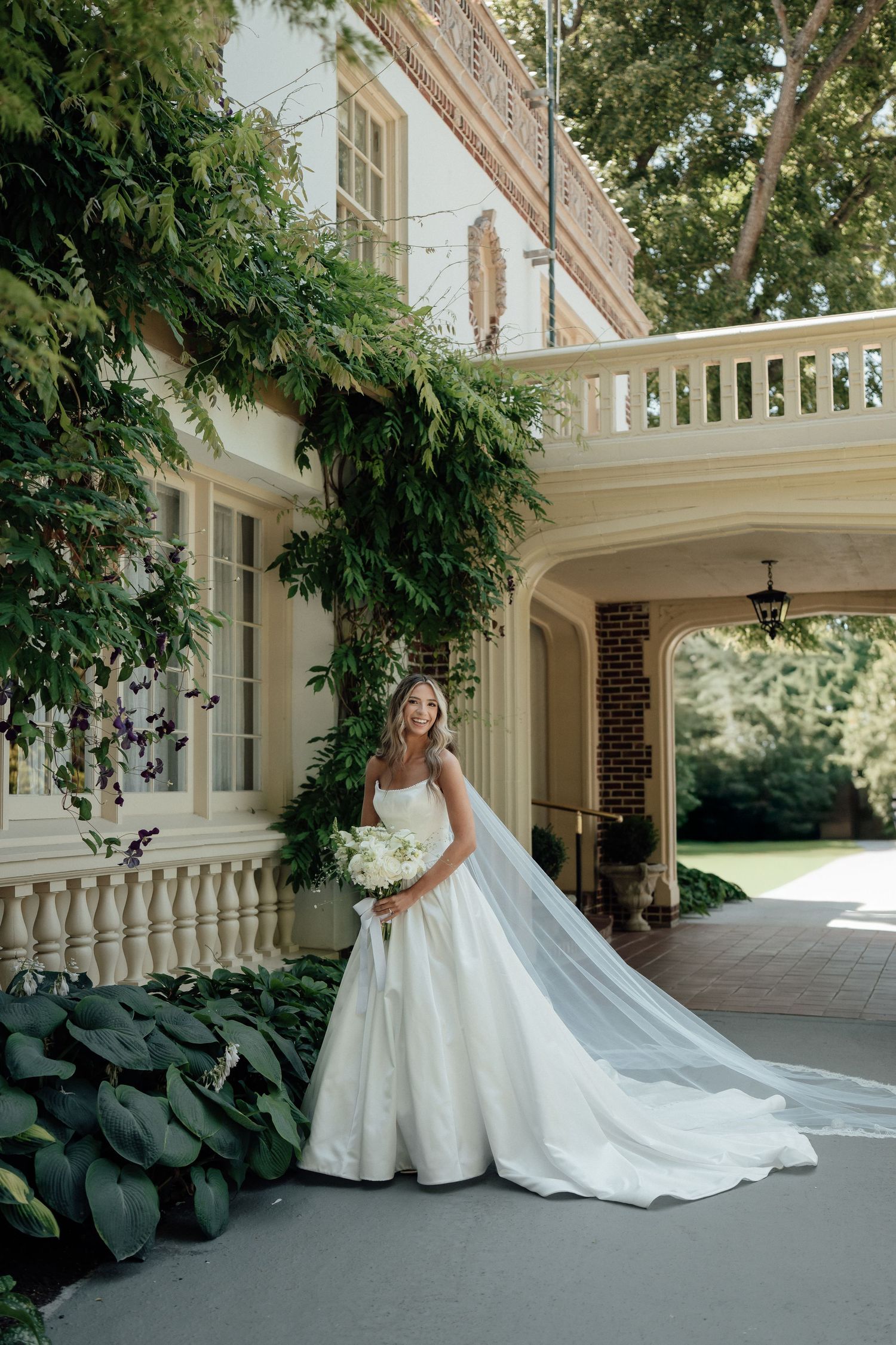 Beautiful wedding dress with long train photographed on elegant porch with white columns and lush greenery.