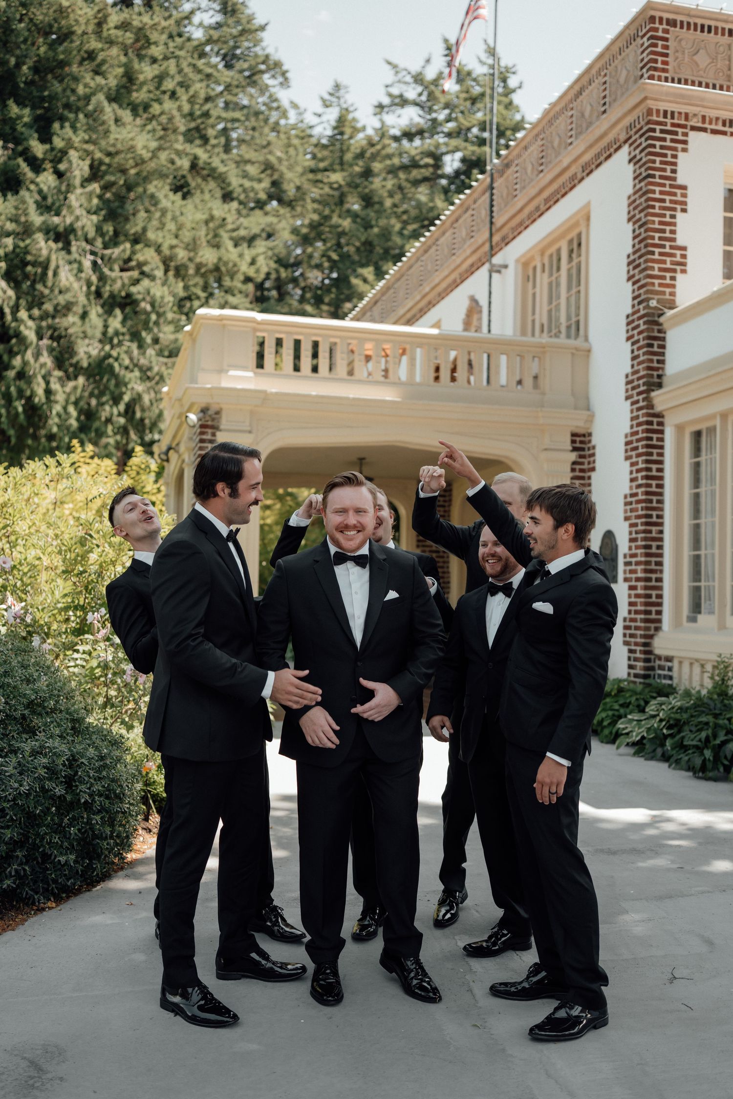 Groomsmen in black tuxedos celebrate and pose together outside a brick building during wedding festivities.