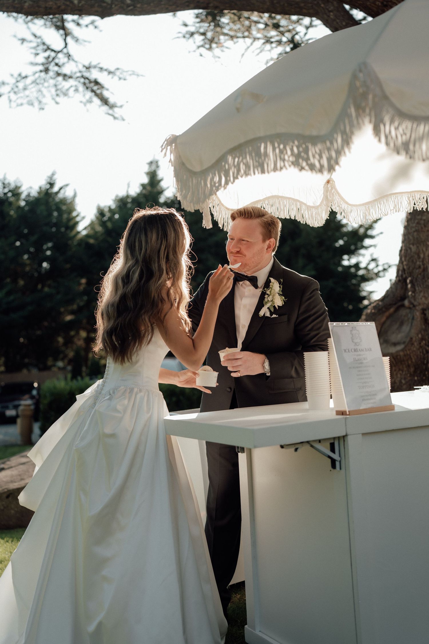 A sweet moment between newlyweds sharing dessert under a white fringed umbrella at an outdoor reception.