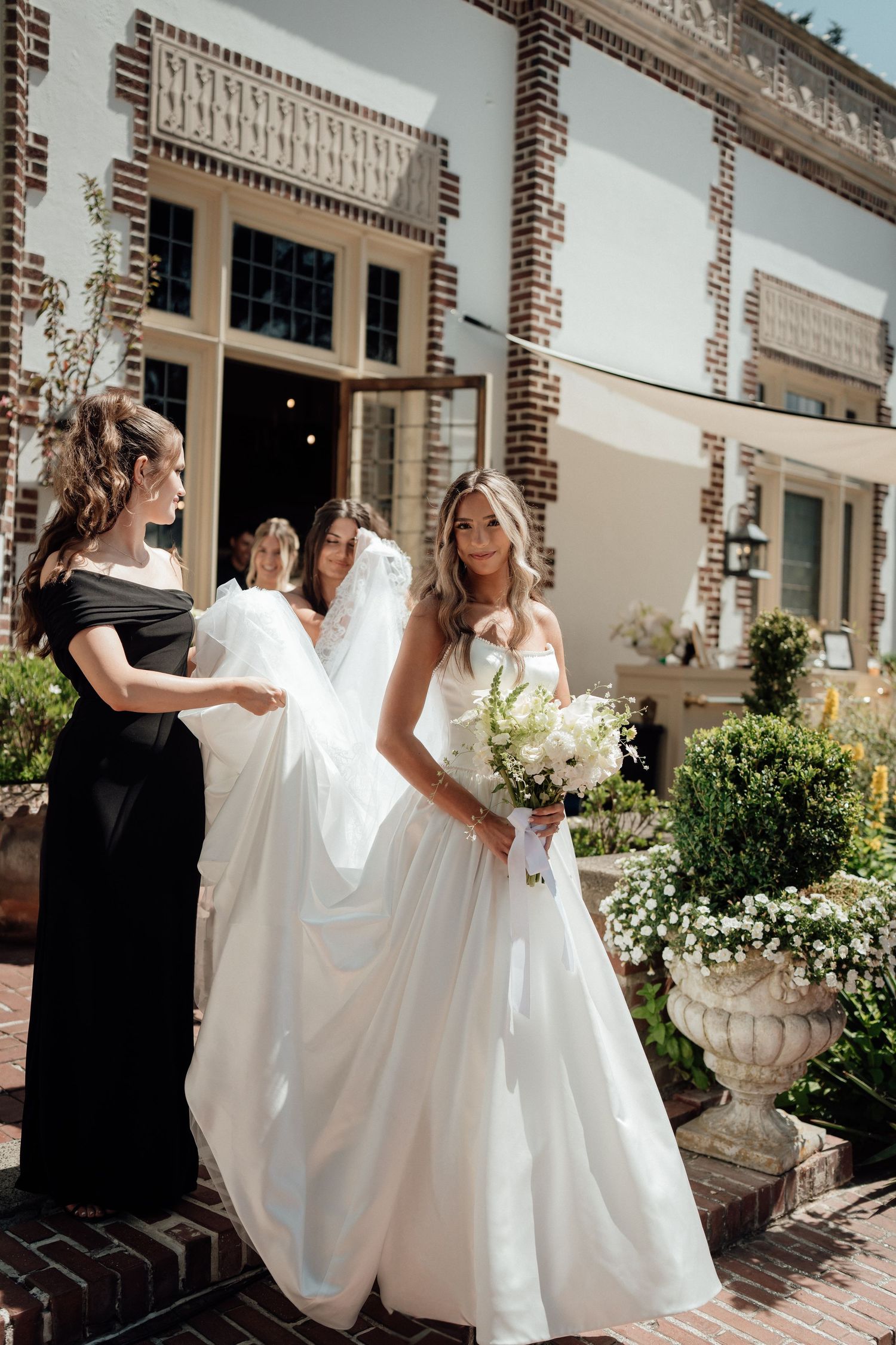 Multiple views of bridesmaids helping to fluff and arrange a bride's flowing white wedding dress train at a brick venue.