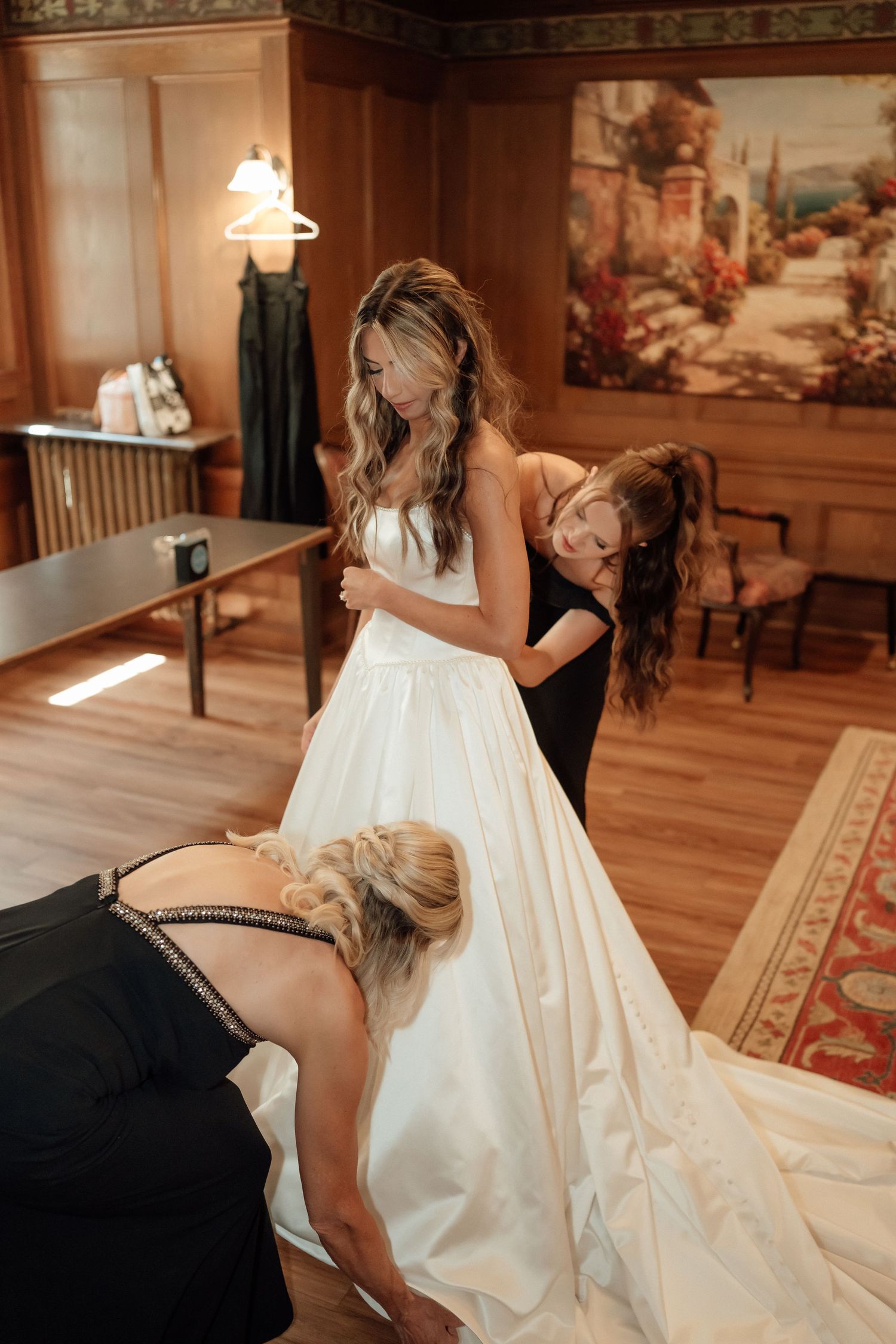 Assistants help a bride with her flowing white wedding gown in an elegant wood-paneled room.