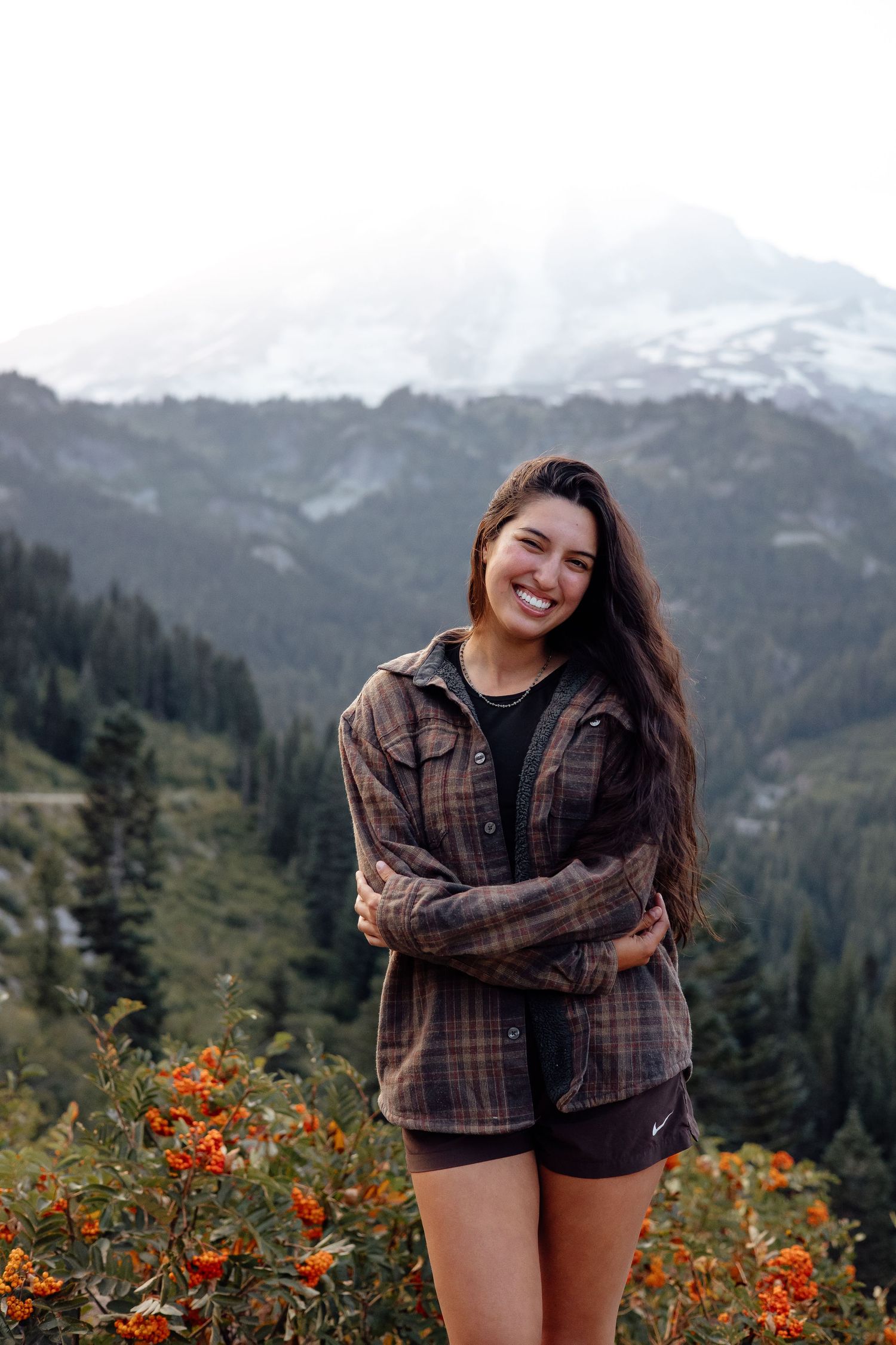 Hiker wearing plaid flannel and shorts stands on mountain trail surrounded by orange wildflowers and snow-capped peaks.