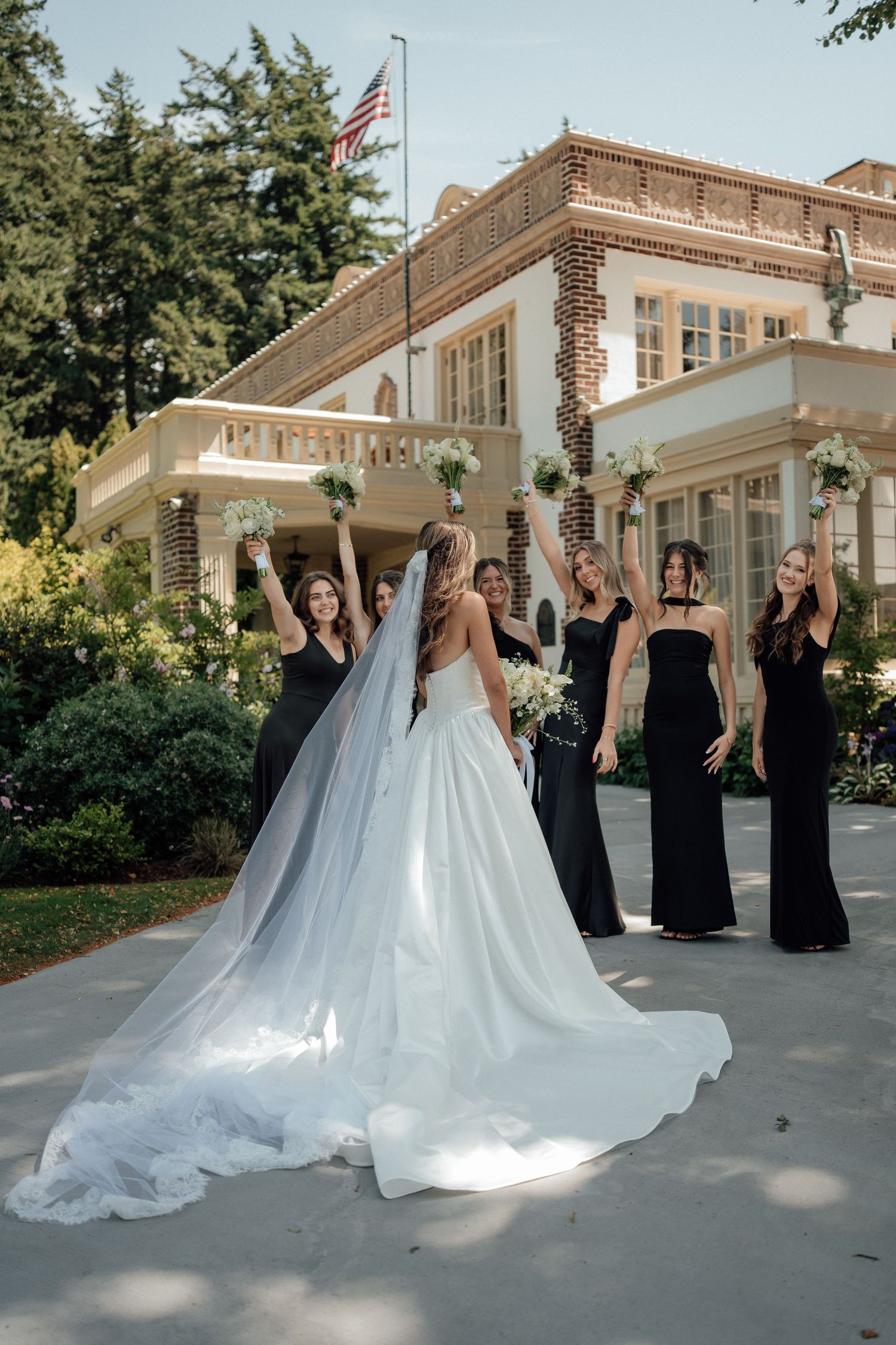 A wedding celebration outside a classic brick building with bridesmaids in black dresses cheering with the bride.
