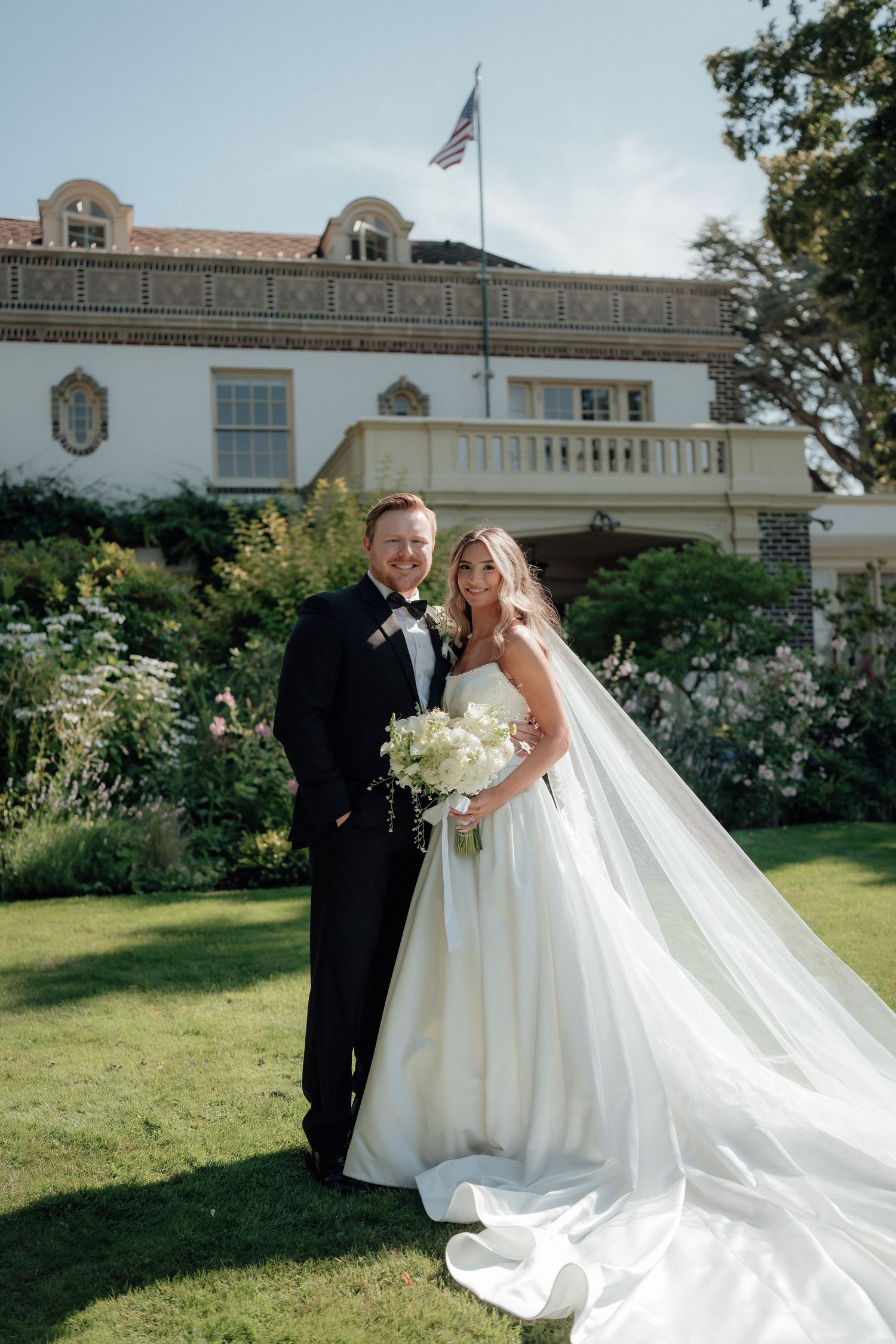 A wedding couple poses on a manicured lawn in front of an elegant white building with an American flag.