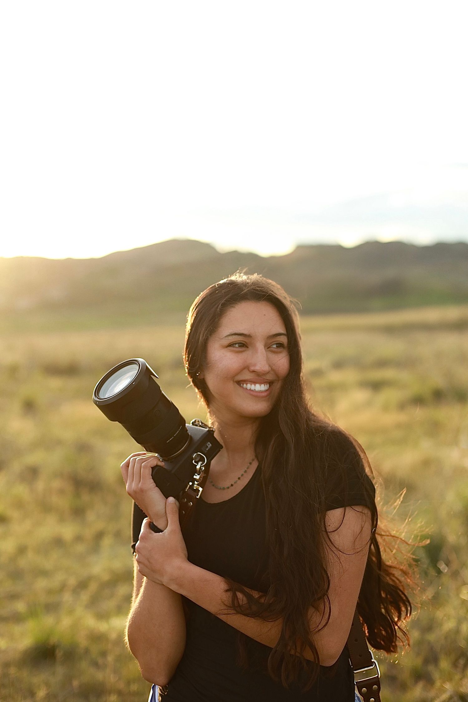 Professional photographer holding camera equipment smiles while standing in golden field during sunset.