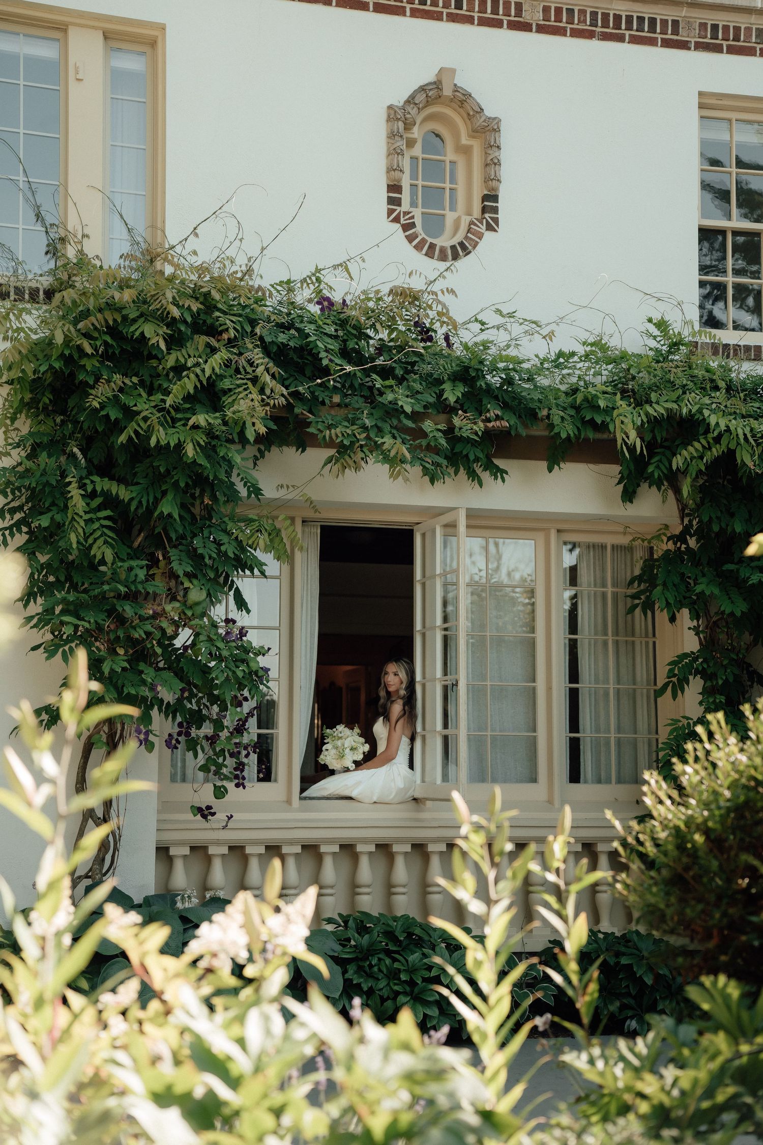 A bride sits elegantly in a window frame of a historic mansion surrounded by lush greenery.