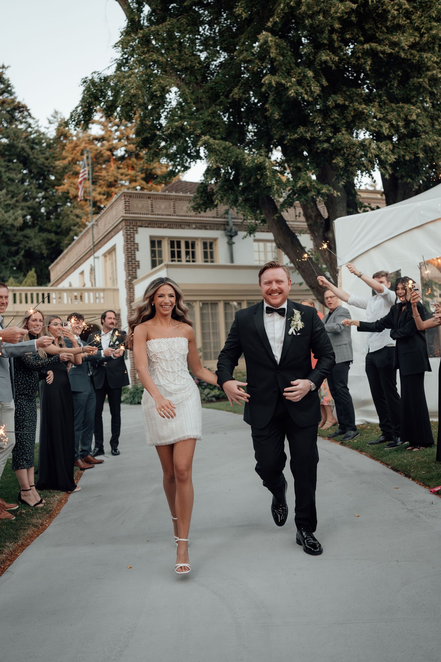 Wedding couple joyfully exiting down a pathway as guests cheer in front of a stately mansion.