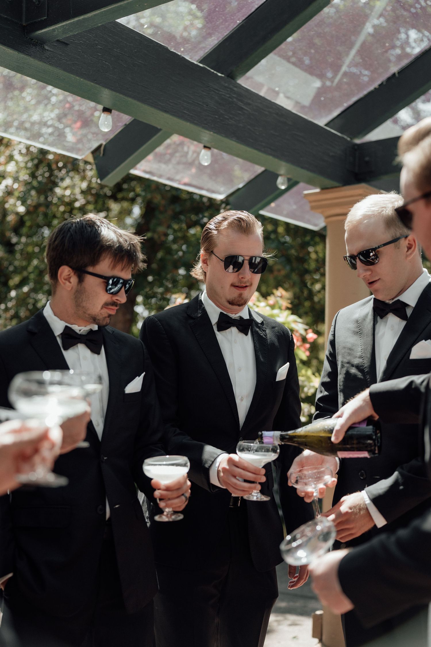 Groomsmen in black tuxedos and sunglasses holding cocktails at an outdoor wedding reception gathering.