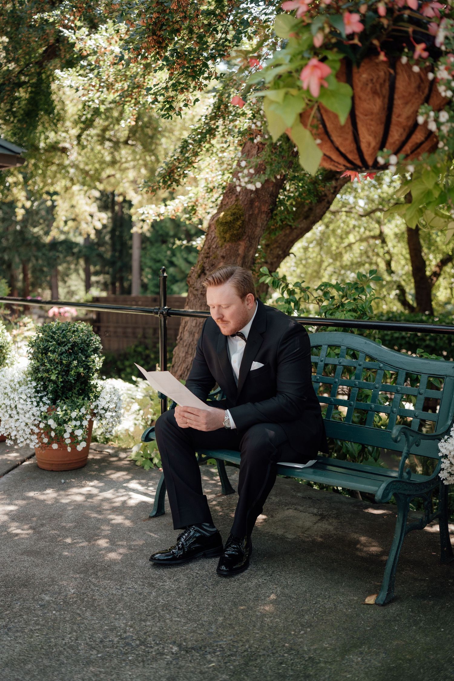 A formal tuxedo-clad figure reading a paper while seated on a green garden bench beneath flowering trees.