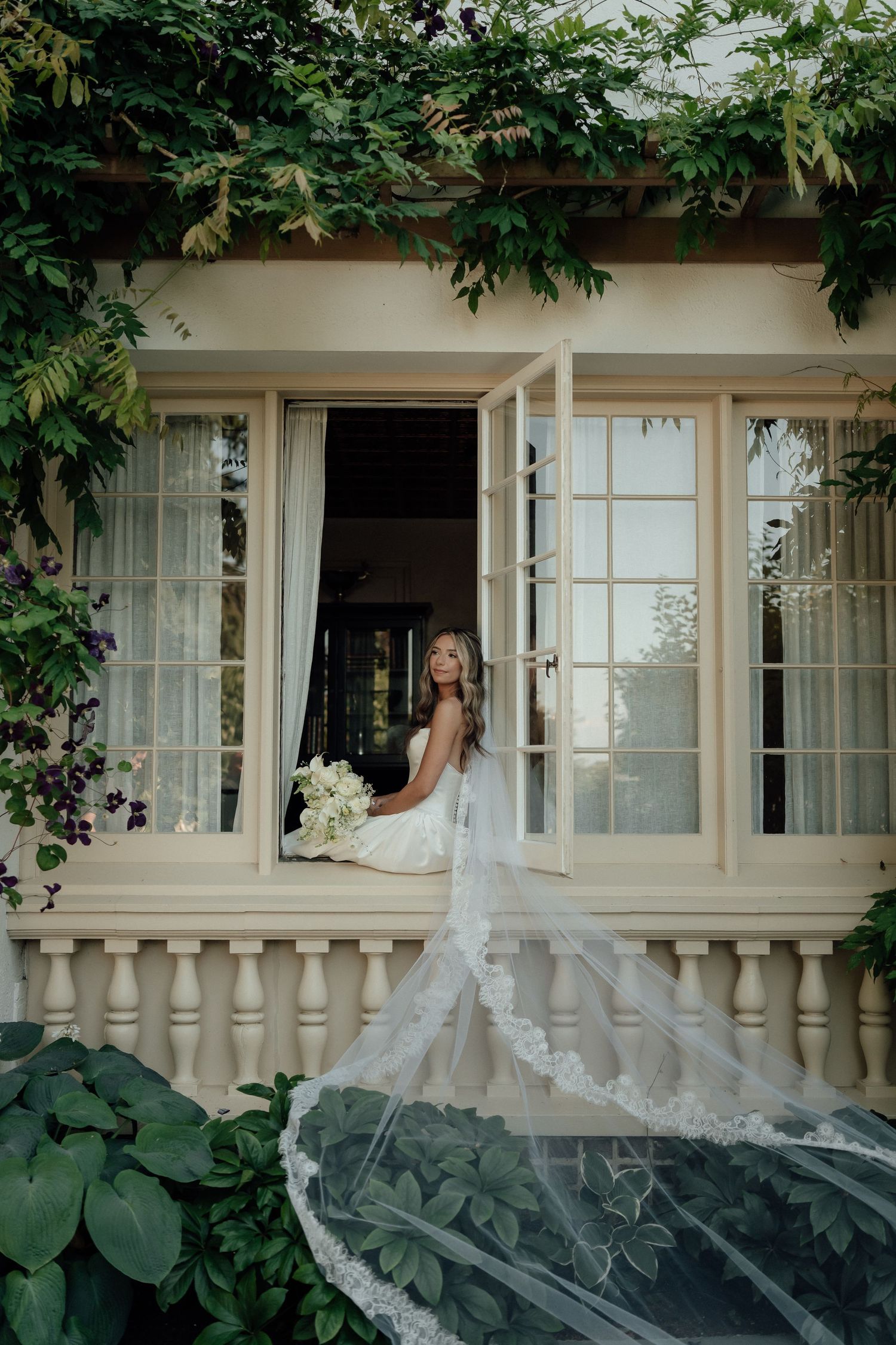 A beautiful bridal portrait through an ornate window with flowing veil and white bouquet amongst lush greenery.