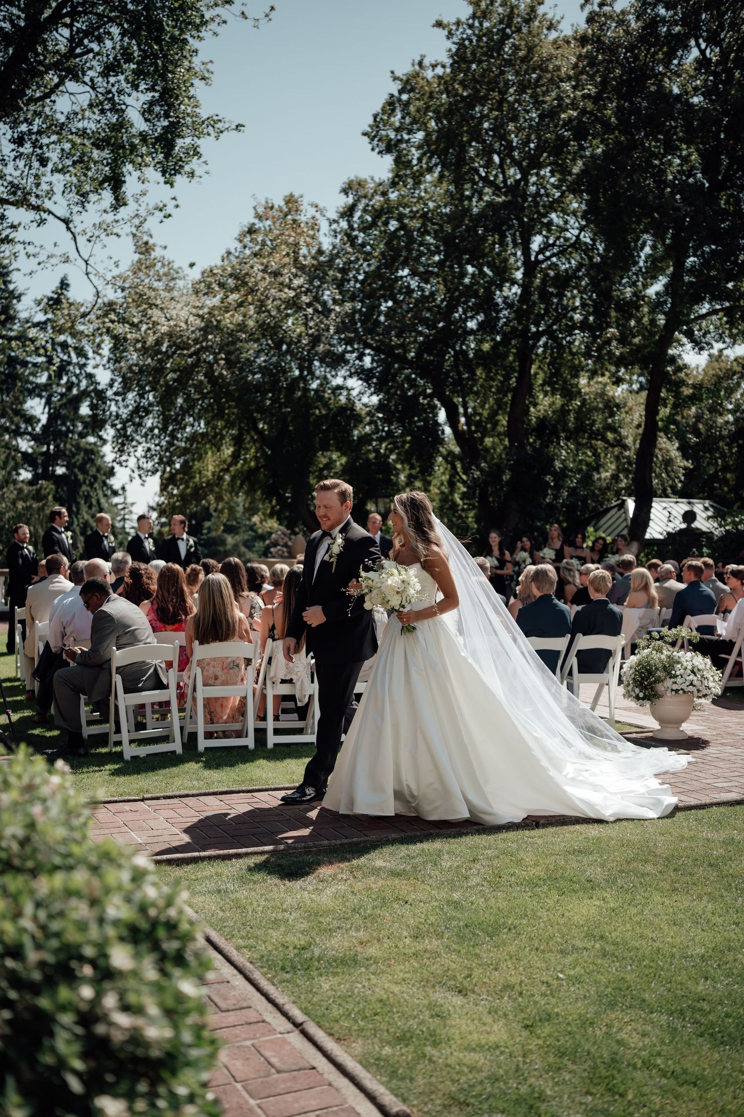 Wedding ceremony takes place outdoors on a beautiful sunny day with white chairs arranged on a lawn for guests.