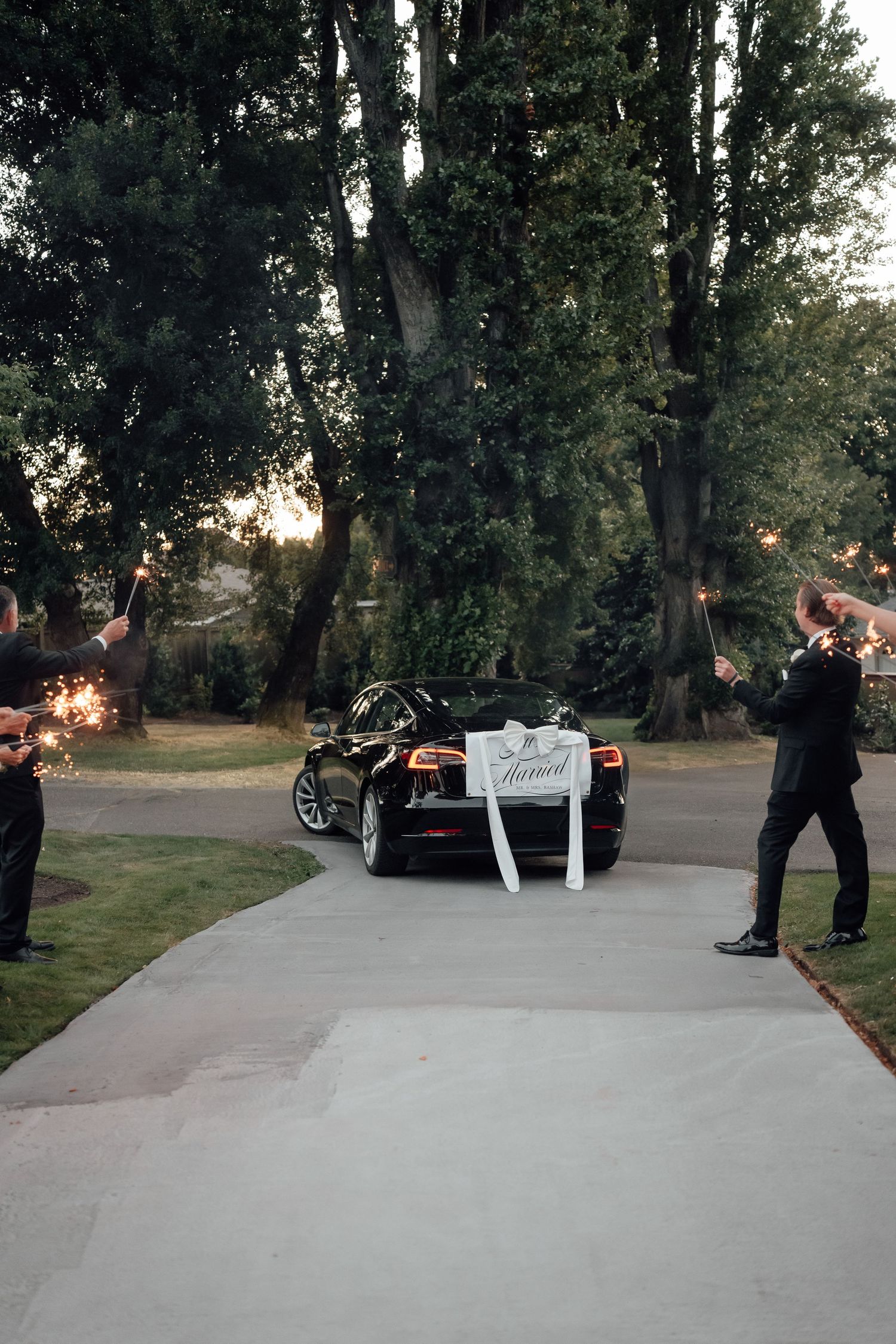 Wedding car decorated with 'Just Married' sign parked on a pathway at dusk with sparklers lighting the scene.
