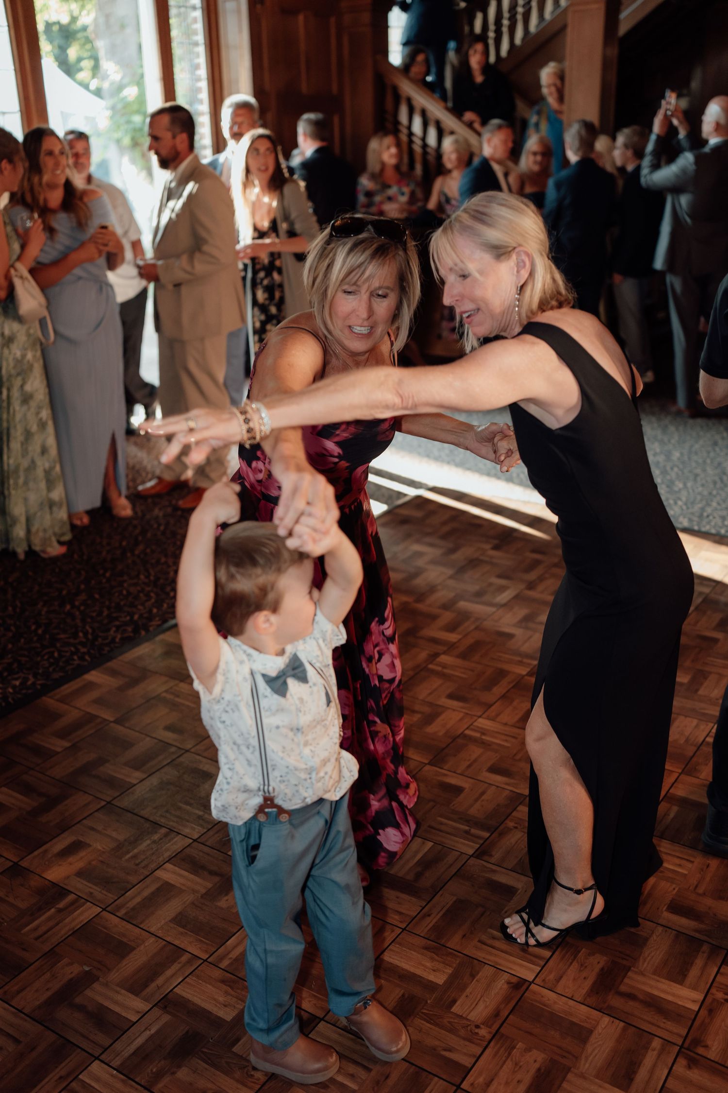 People dance together on a parquet dance floor at a wedding reception while guests mingle in the background.