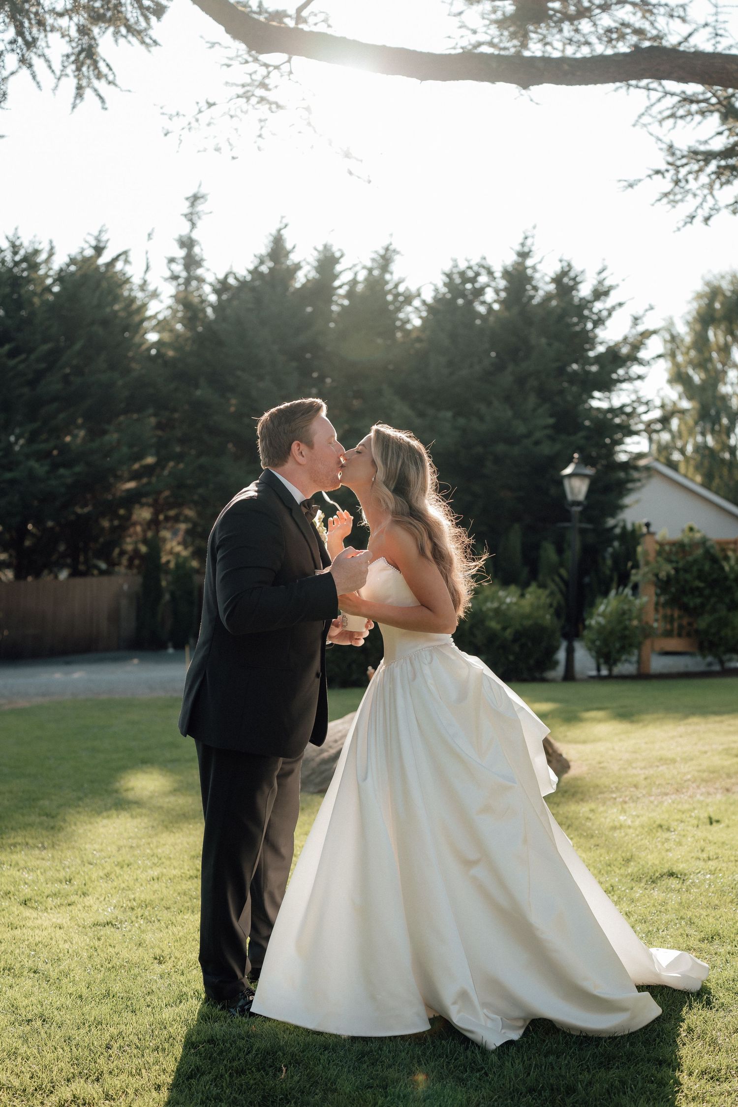 A romantic wedding couple shares an intimate moment on a sunlit lawn at dusk.