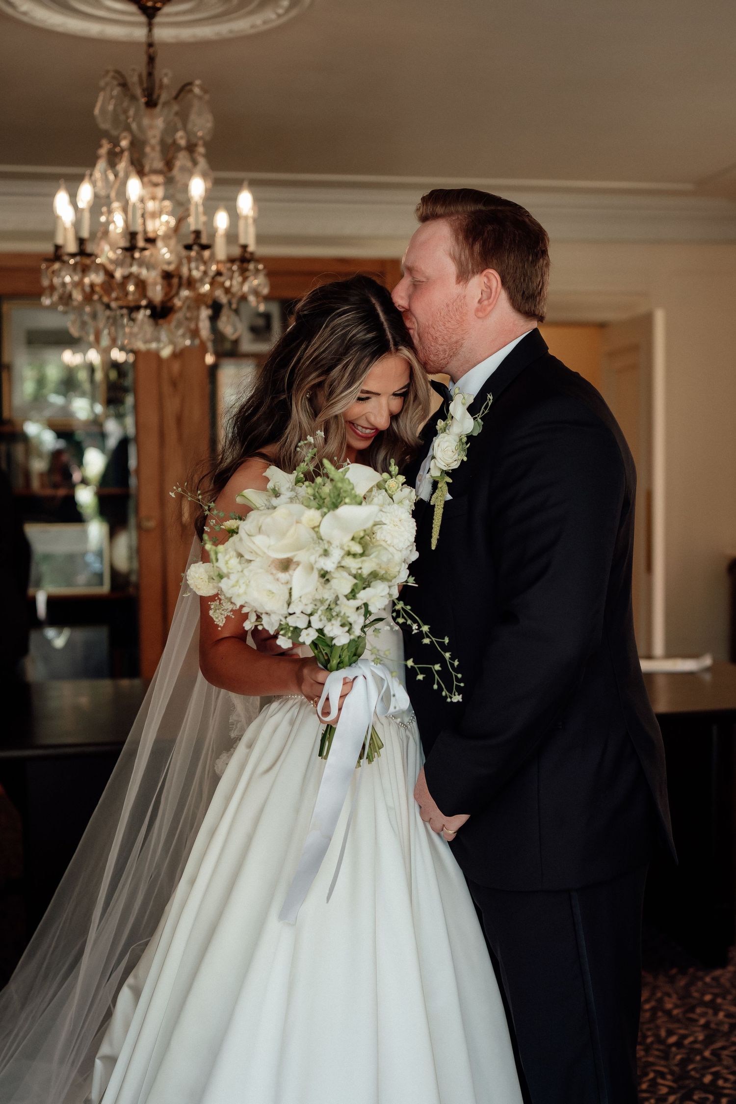 Wedding couple shares an intimate moment under a crystal chandelier with white floral bouquet.