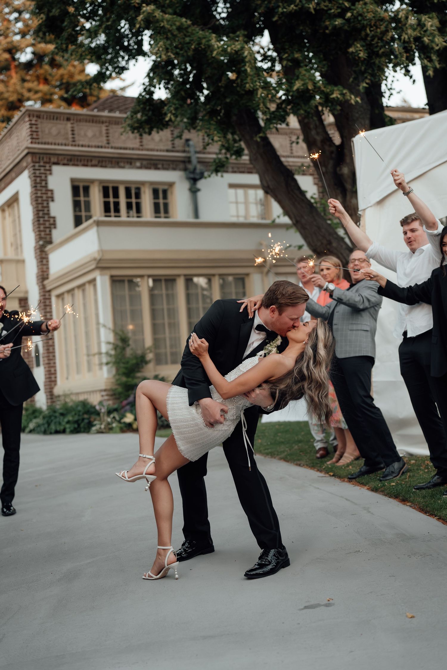A romantic dip dance move at a wedding with guests cheering in the background near an elegant brick building.
