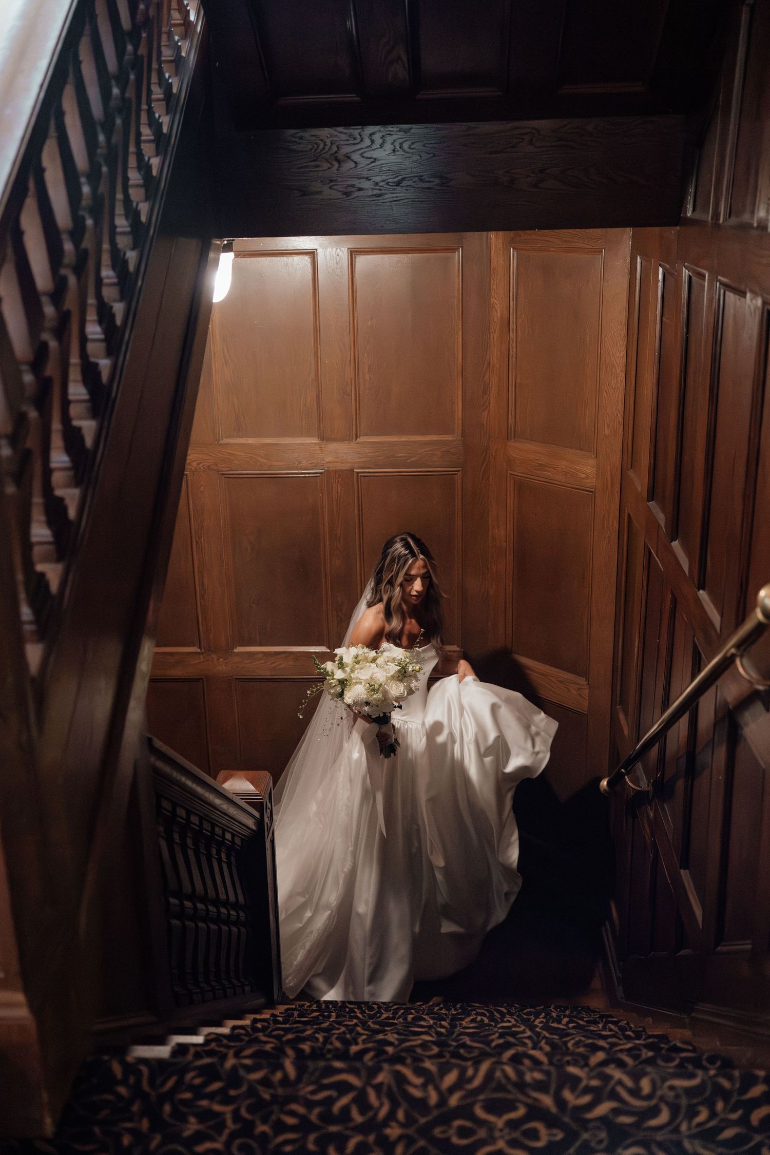 A dramatic wedding photo of a bride in a flowing white dress descending a wood-paneled staircase.