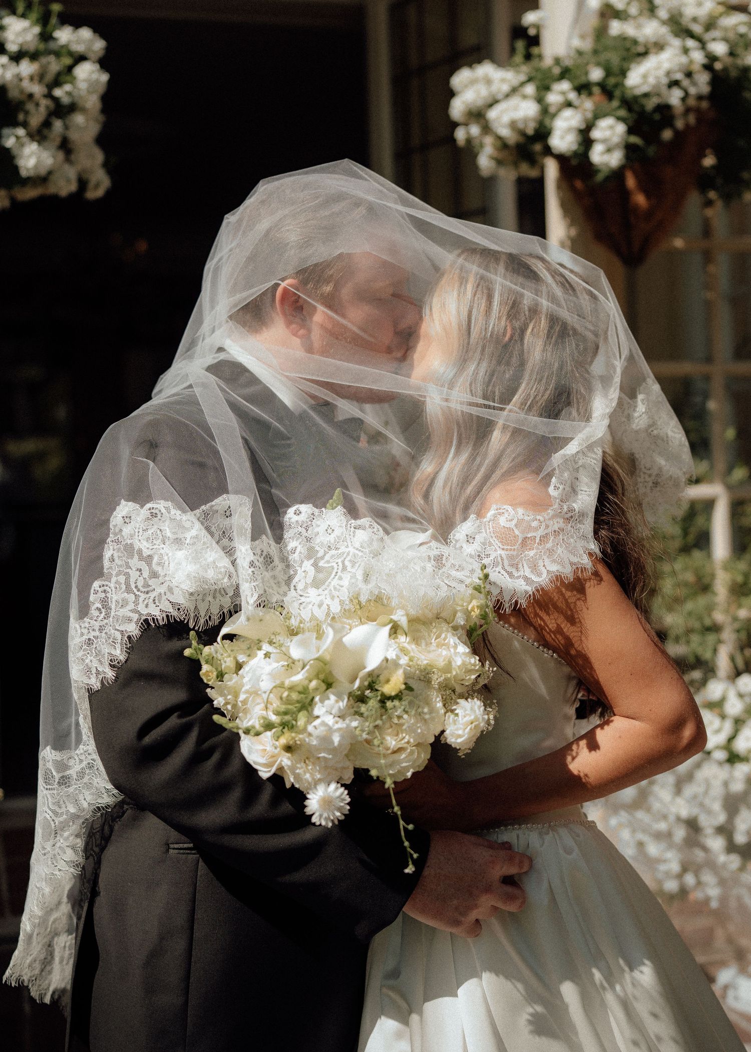 Romantic wedding portrait with couple embracing under lace veil surrounded by white flowers.