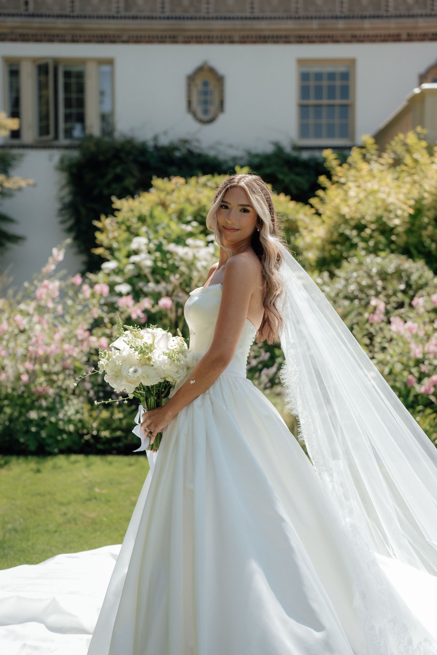 A beautiful wedding gown with flowing veil photographed in a sunny garden with white flowers and classic architecture.