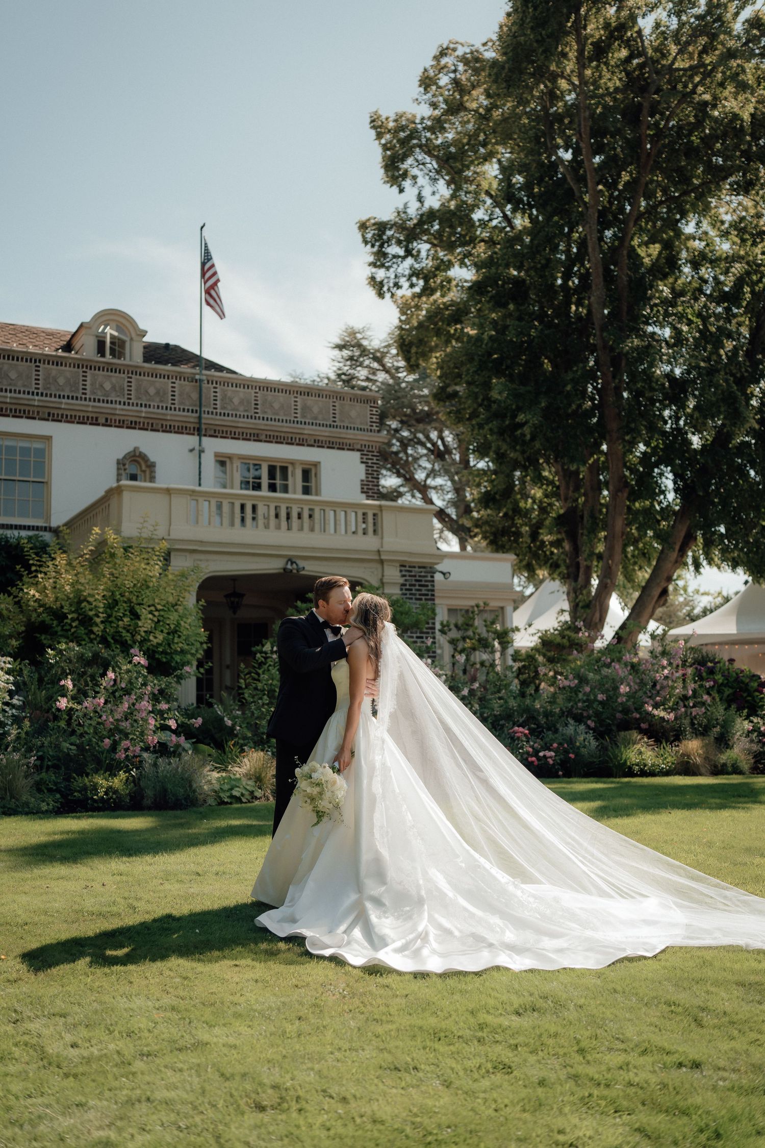 A romantic wedding couple embracing on a manicured lawn in front of a grand estate with an American flag.