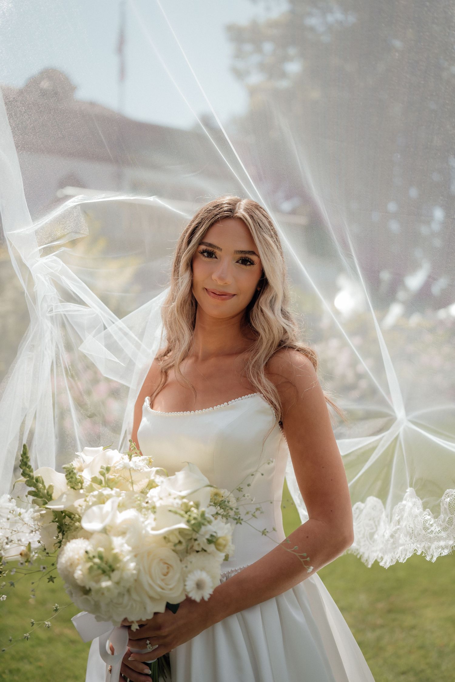 A bride wearing a white wedding dress holds a bouquet of white flowers under a flowing veil in an outdoor setting.