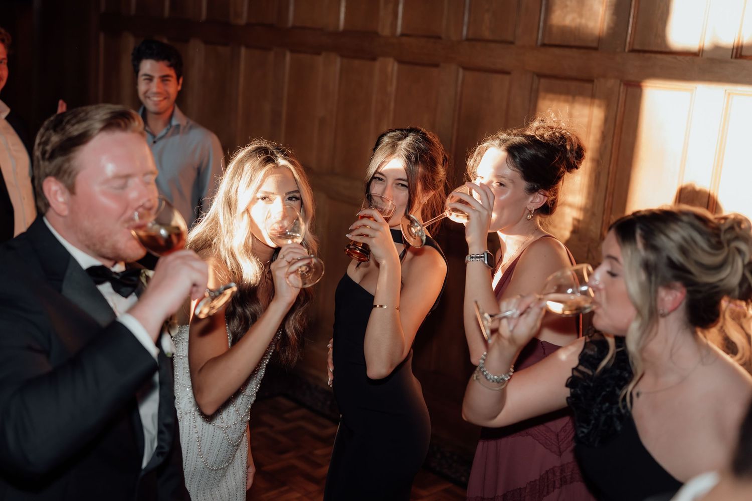 Wedding guests enjoying drinks and laughter at an evening reception in a wood-paneled venue.