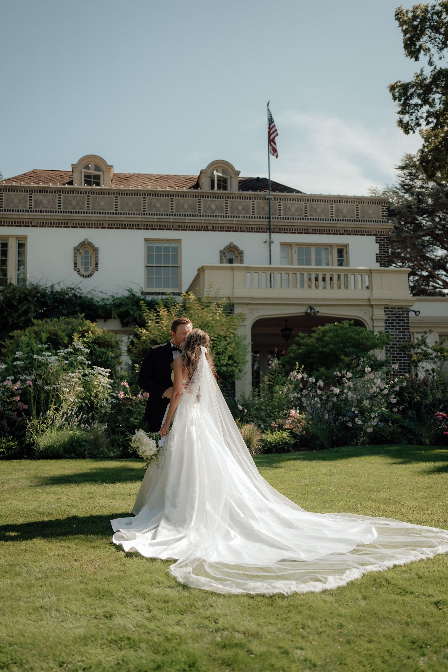 A wedding couple embraces on a manicured lawn in front of an elegant white mansion with an American flag.