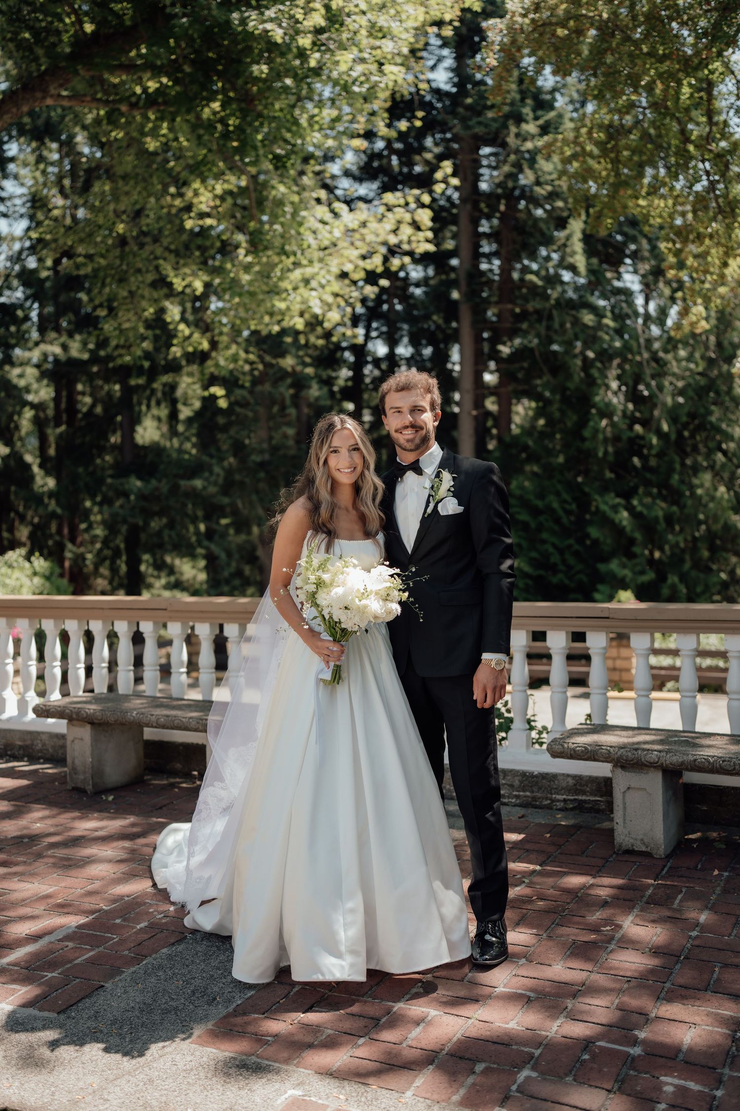 A wedding couple poses for a portrait on a brick terrace surrounded by lush greenery.