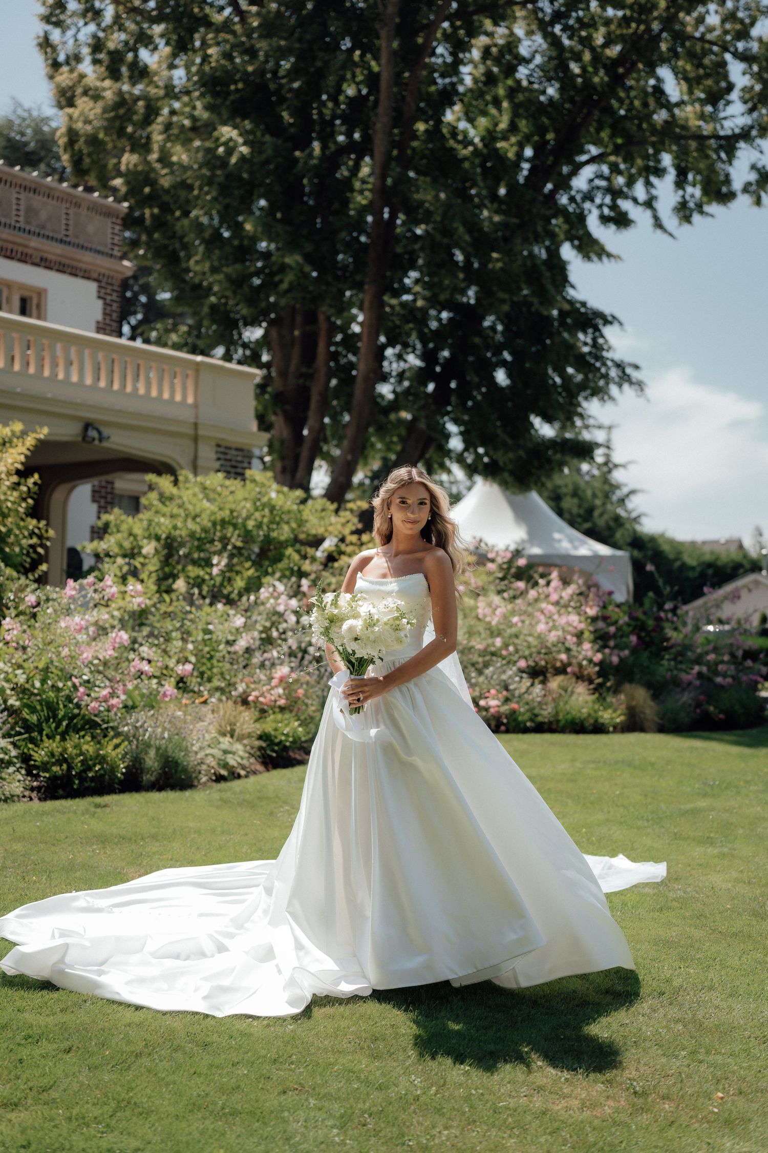 Bride in flowing white gown stands on green lawn with garden and historic mansion in background.