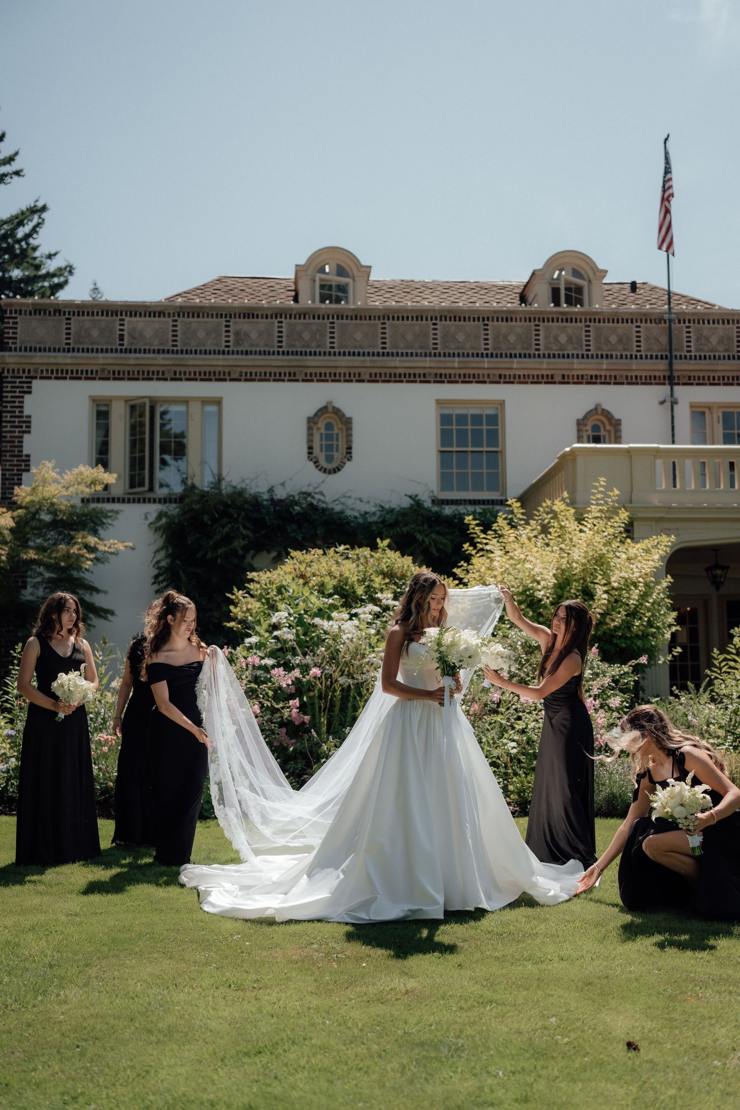 Bridal party arranging a wedding dress train on sunny lawn in front of elegant Spanish-style building with American flag.
