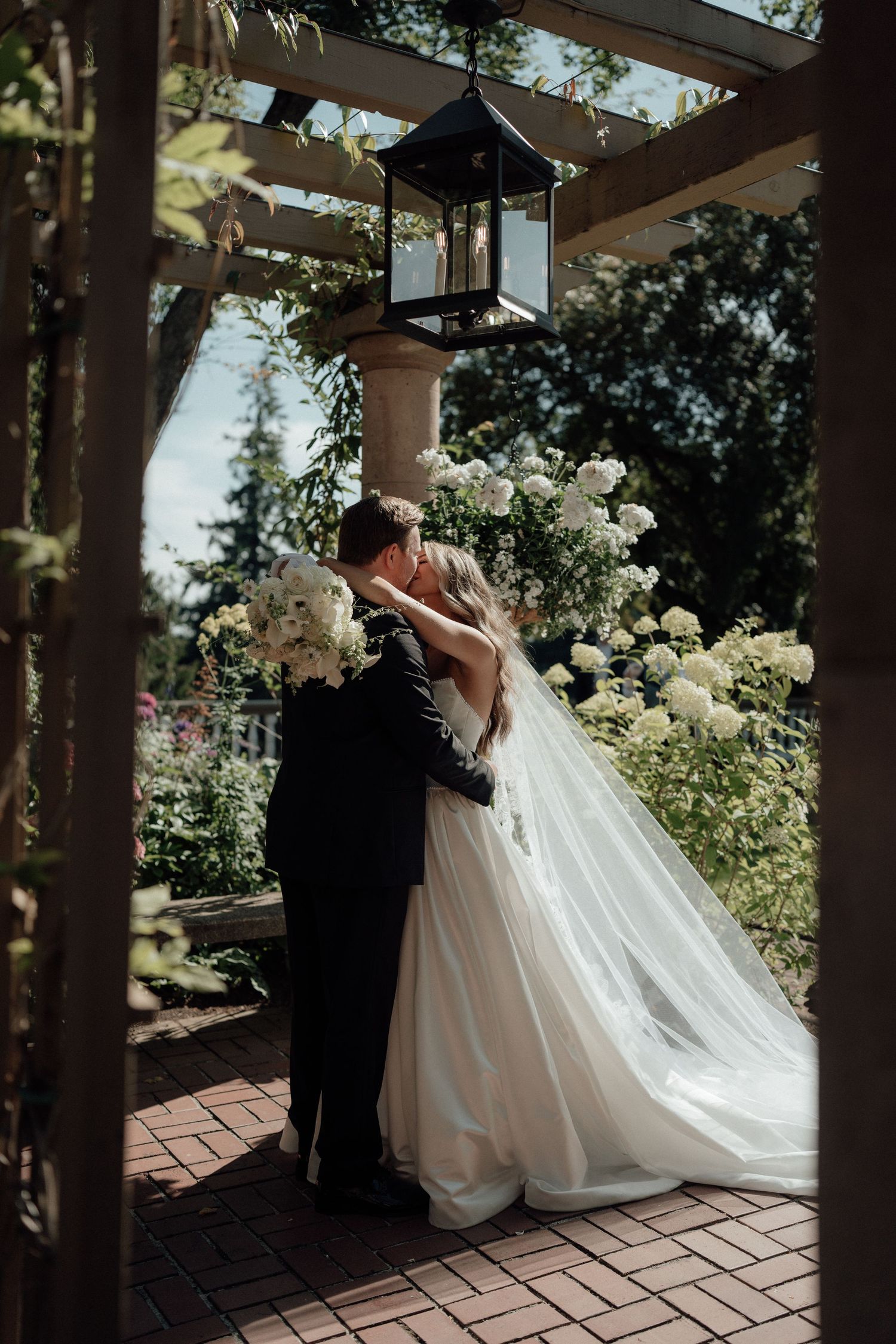 A romantic wedding couple shares a kiss under a lantern-lit pergola surrounded by white flowers.