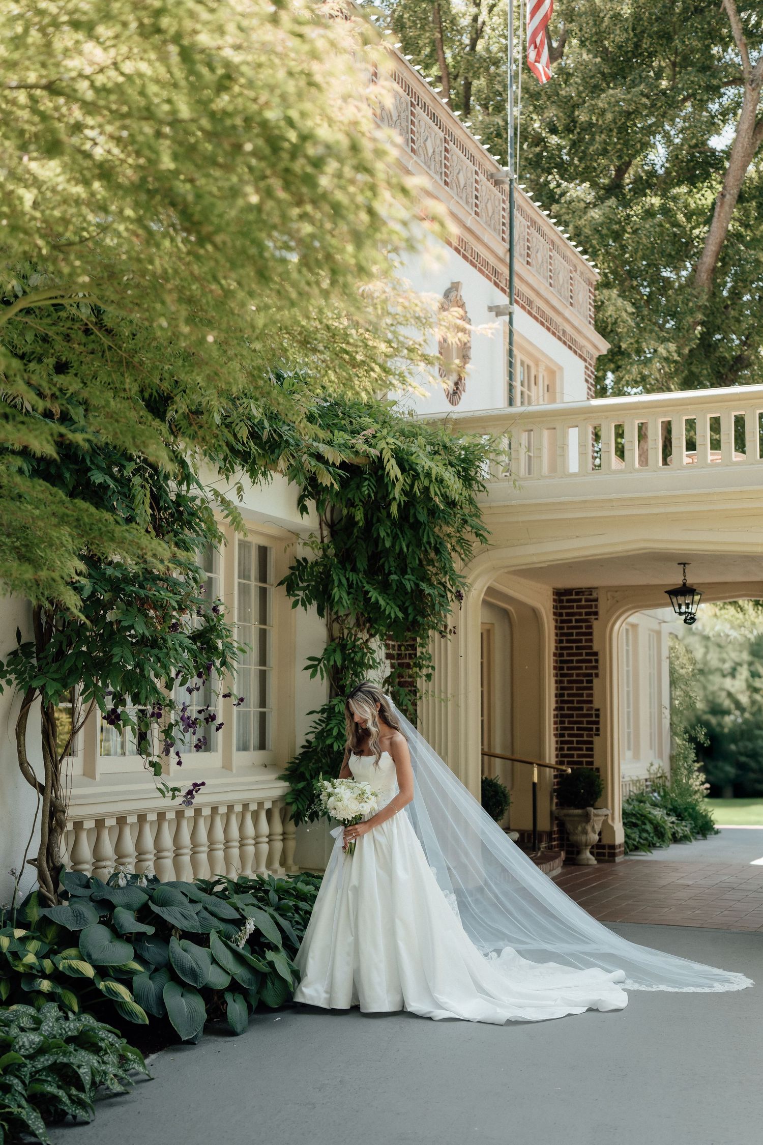A bride in a white gown with long train poses against an elegant white mansion with climbing vines.