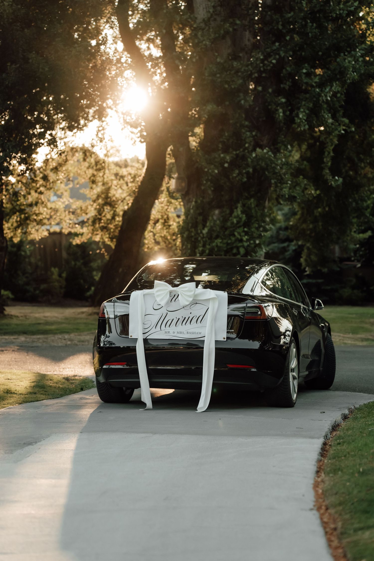 A black car decorated with 'Just Married' sign and ribbons photographed at sunset on a winding path.