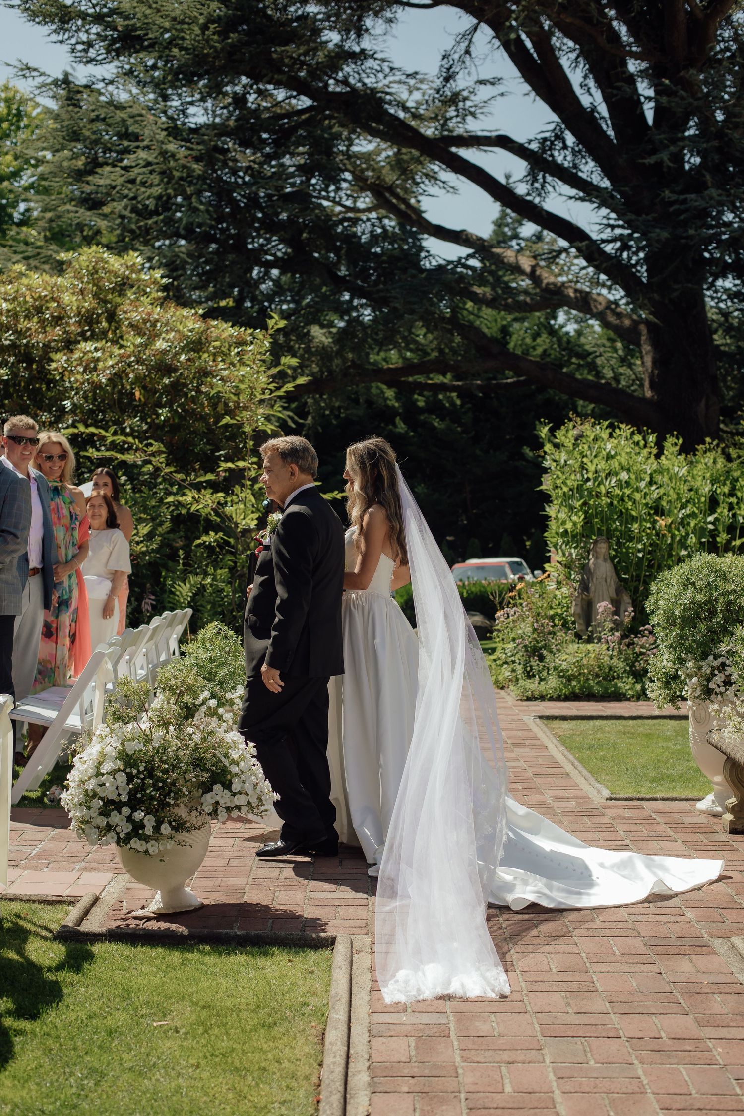 Wedding ceremony on a brick path with white chairs and lush garden landscaping.
