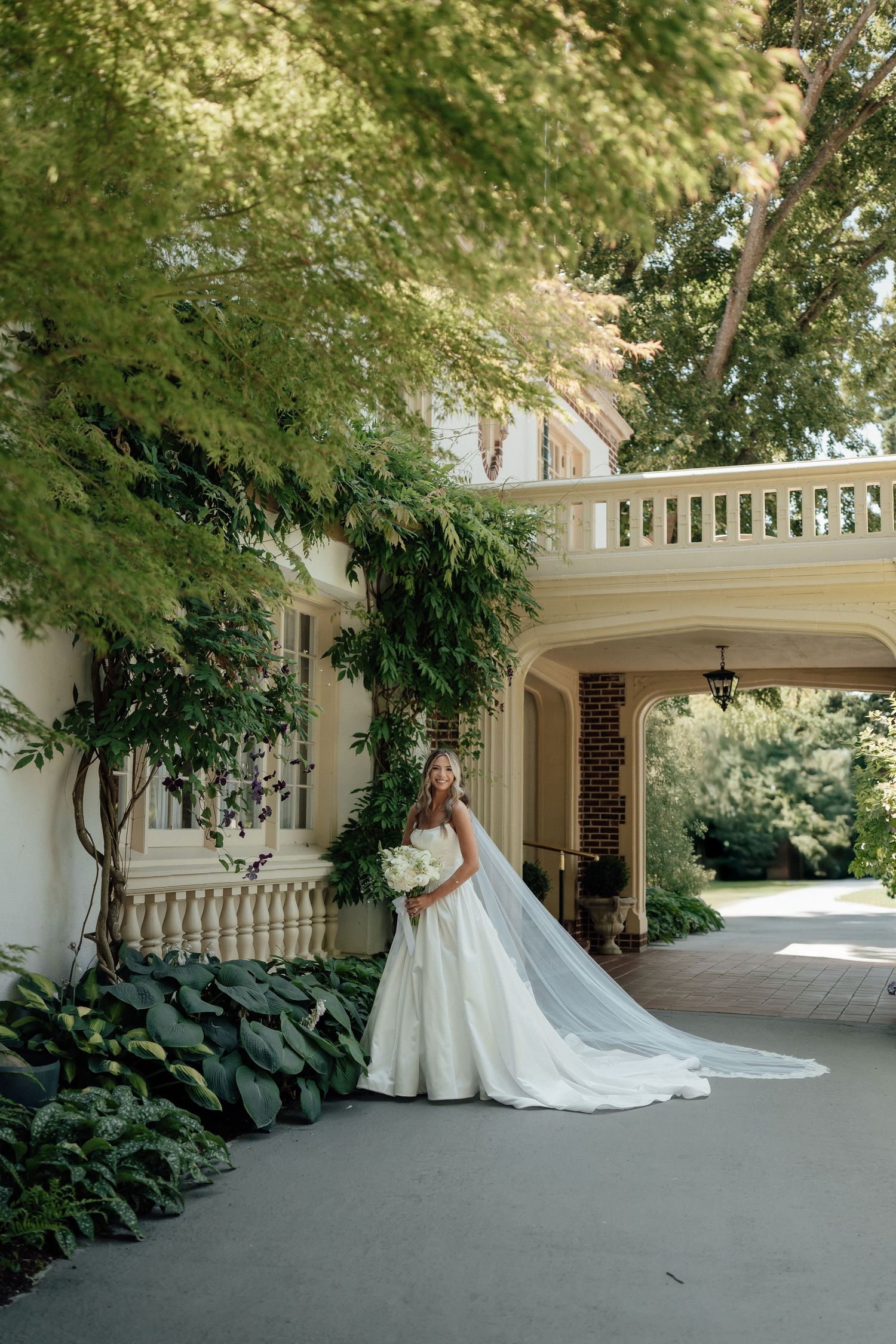 A bride in a flowing white gown stands gracefully under an arched portico surrounded by lush greenery.