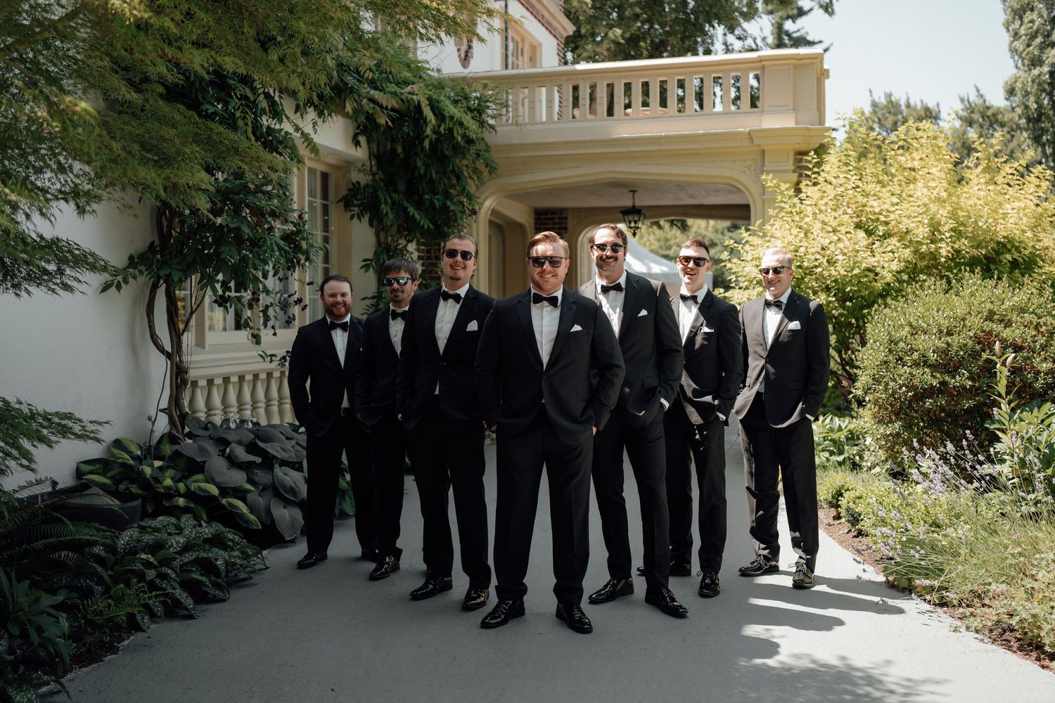 Groomsmen in black tuxedos stand in line on a garden terrace of an elegant mansion.