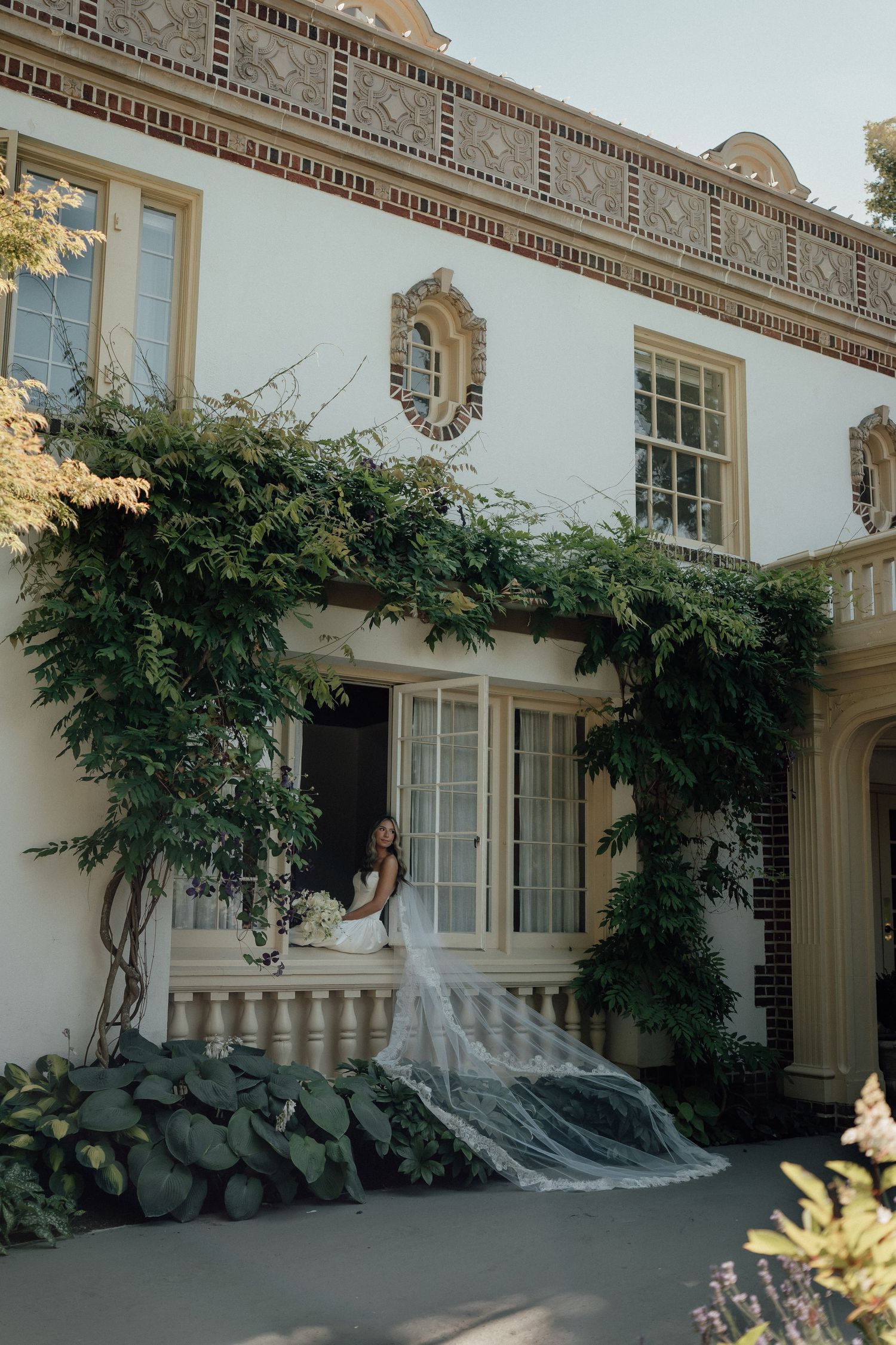 A bride poses in an architectural window frame with flowing veil against a classic white mansion facade.