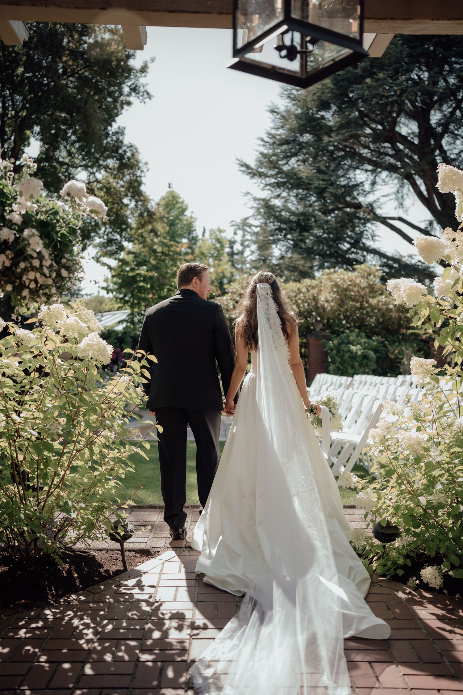 Romantic wedding couple walking hand in hand through garden pathway under hanging lantern.