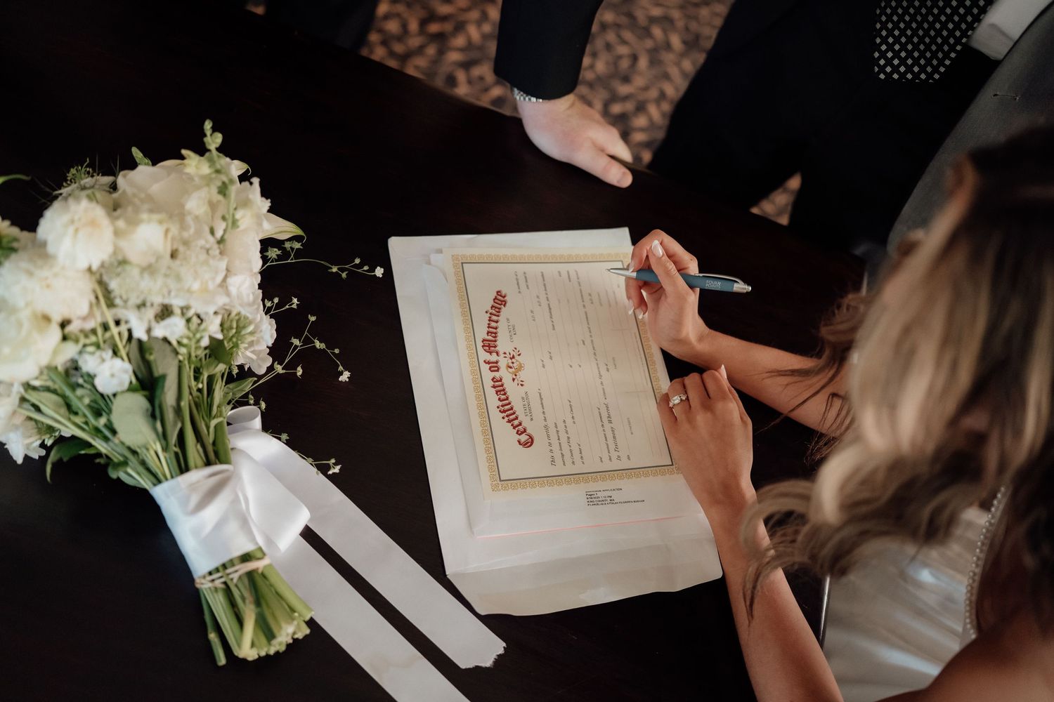 Hands signing a wedding certificate with a white floral bouquet on a black surface.