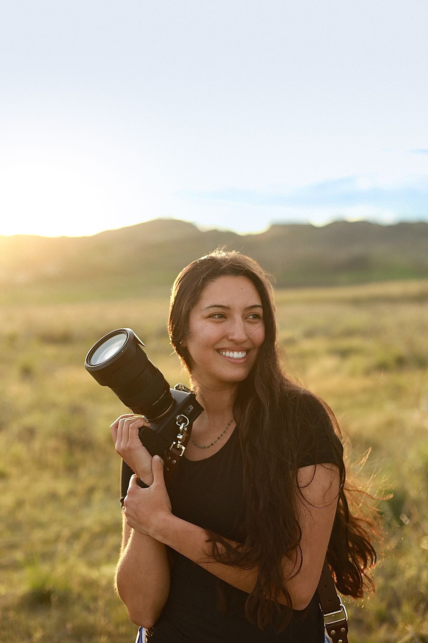 Professional photographer holding camera equipment while photographing outdoors in golden sunlight at mountainous landscape.