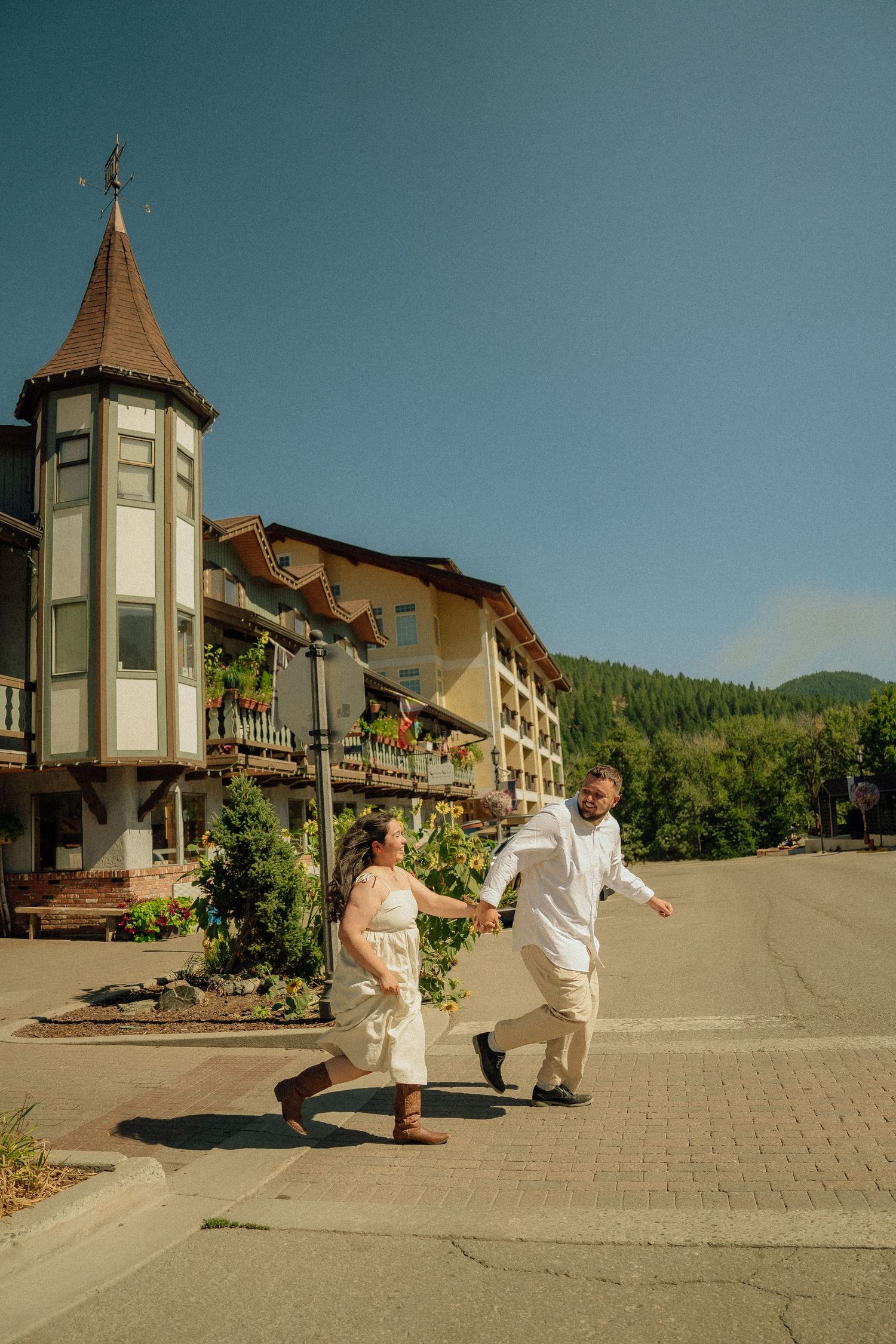 A European-style building with a distinctive turret stands as two people in casual attire walk along a paved pathway.