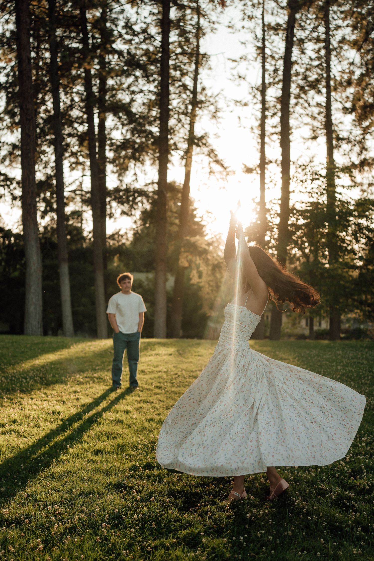 A couple in a sunlit forest setting during golden hour, with a flowing white dress twirling against tall evergreen trees.