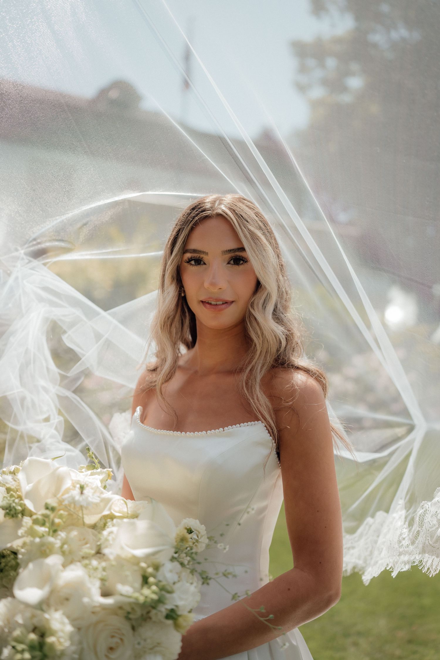 A bride in a white strapless gown with flowing veil poses outdoors surrounded by white floral arrangements.