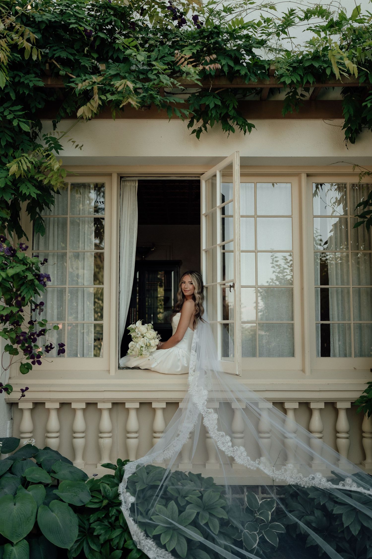 White bridal veil with delicate lace details flows gracefully against column-lined veranda with climbing vines.