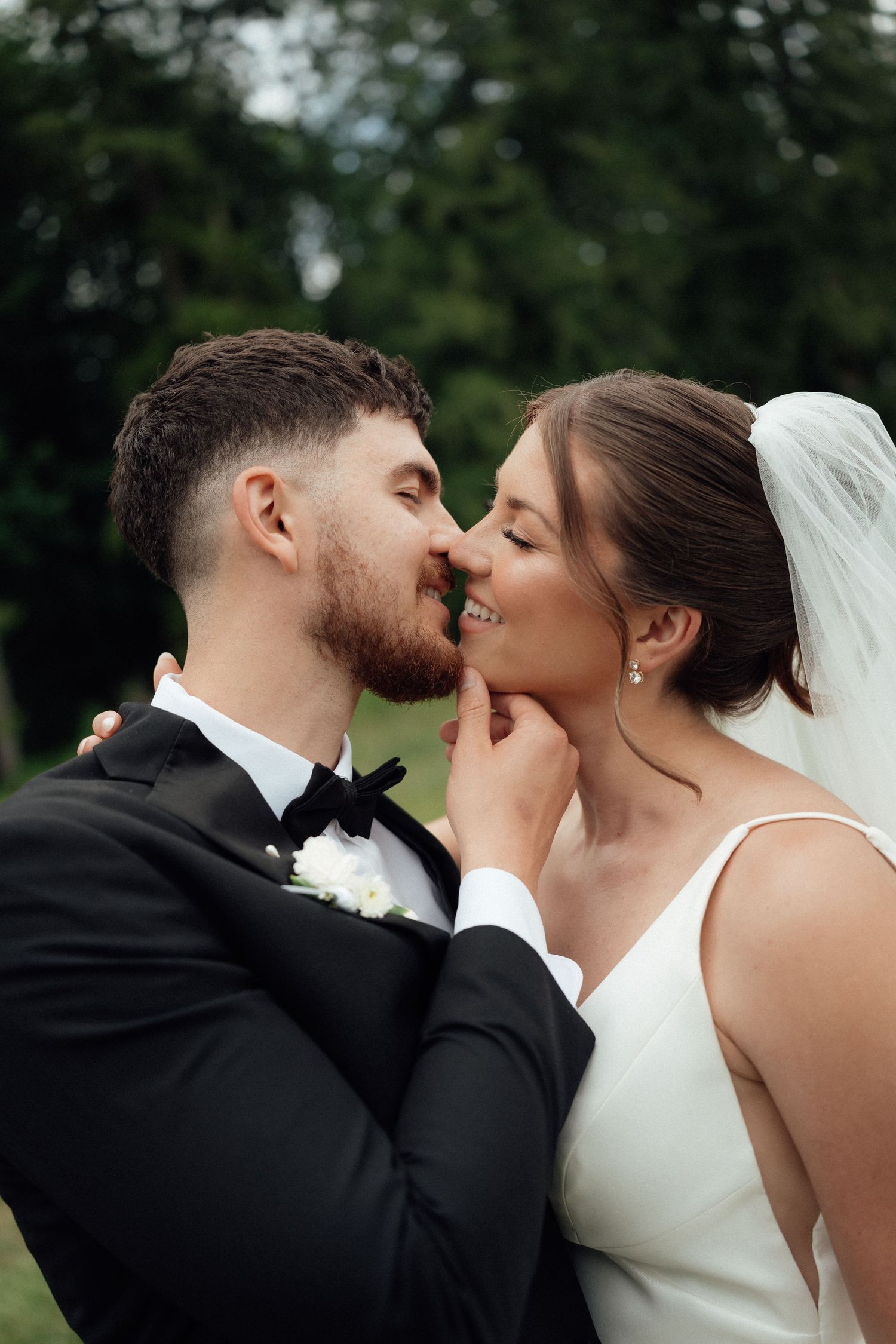 A bride and groom share a romantic almost-kiss moment outdoors, dressed in a white gown and black tuxedo.