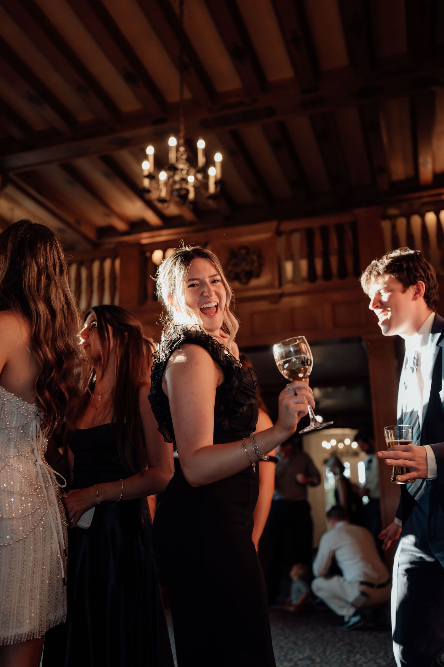 Party guests in formal attire celebrate with wine glasses at an elegant reception with wood beams and chandelier overhead.