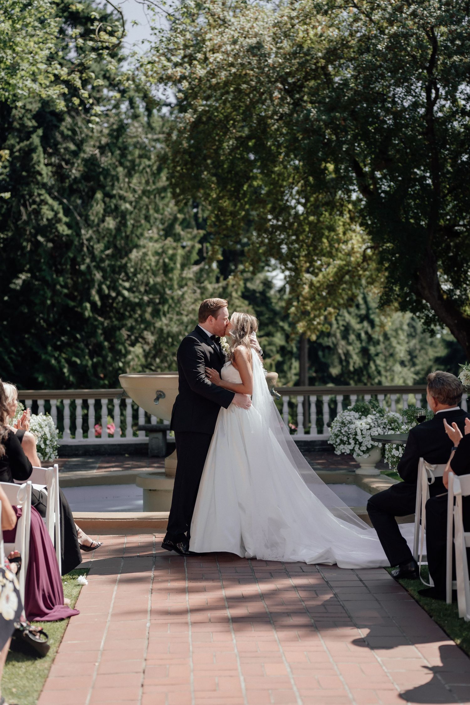 Newlyweds share their first kiss during outdoor garden ceremony surrounded by guests and greenery.