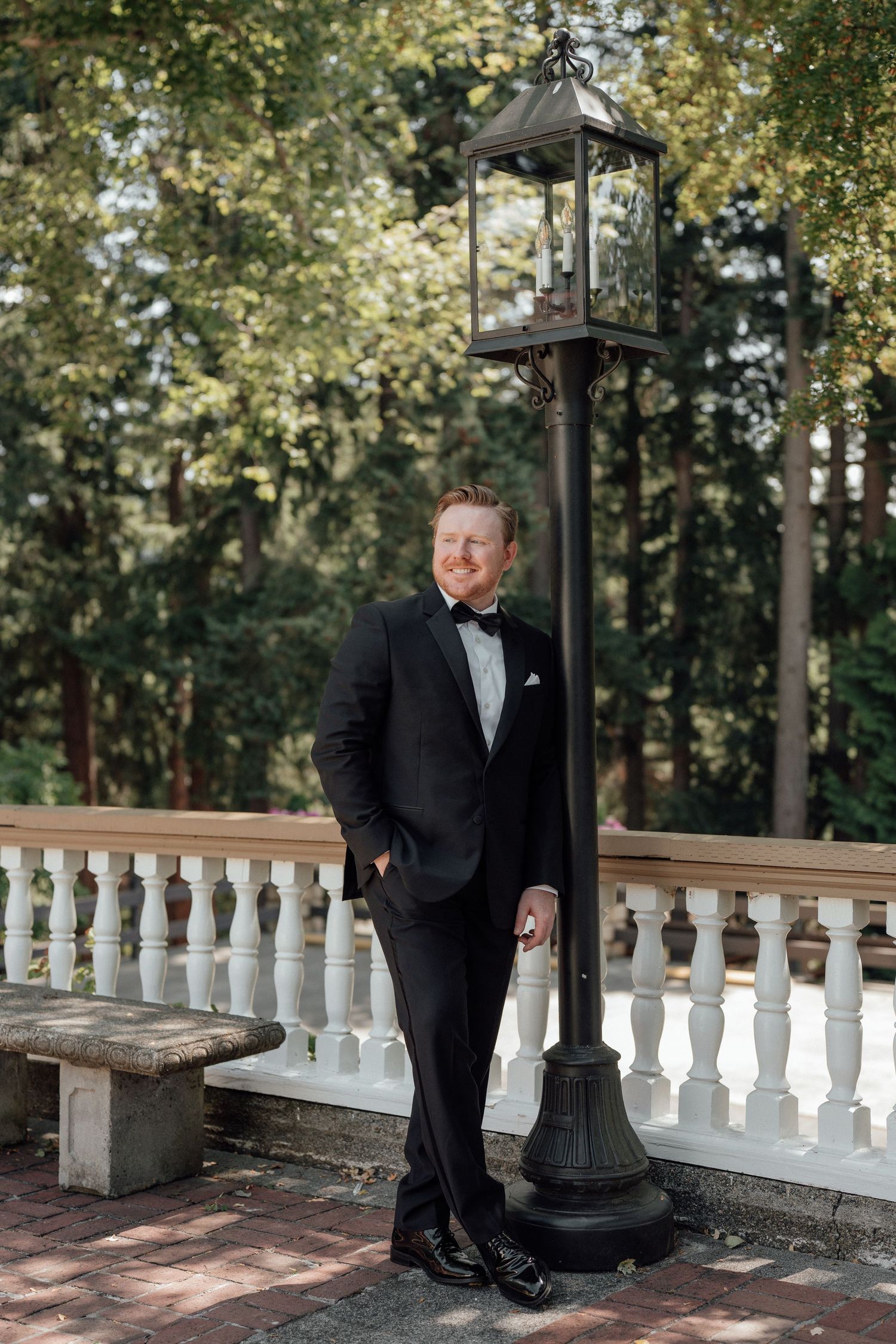 A person in a classic black tuxedo poses elegantly against a vintage lamppost on a white-railed terrace.