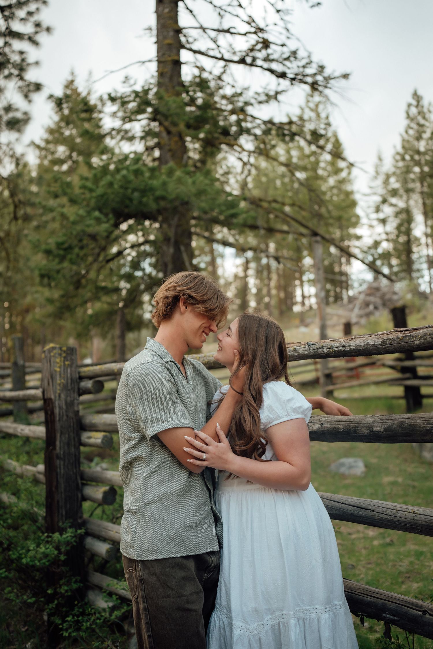 A romantic couple embrace by a rustic wooden fence in a forest setting during golden hour.
