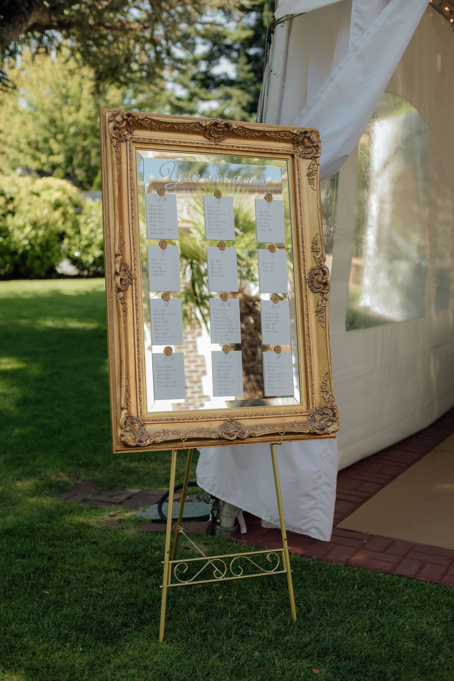 An elegant gold framed seating chart display on an easel for a wedding reception outdoors.